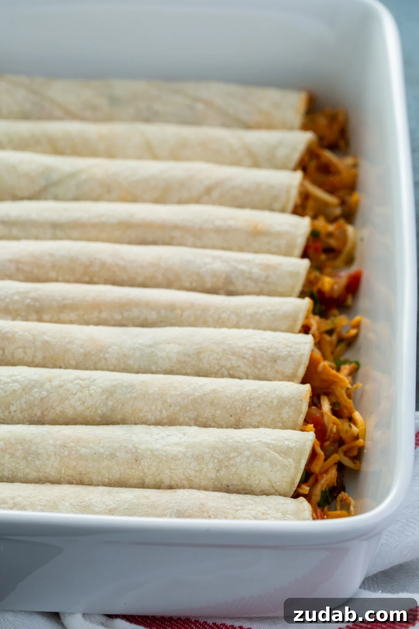 Red Chicken Enchiladas A baking dish filled with uncooked, rolled enchiladas. You can see the chicken filling spilling out the ends of the tortillas.