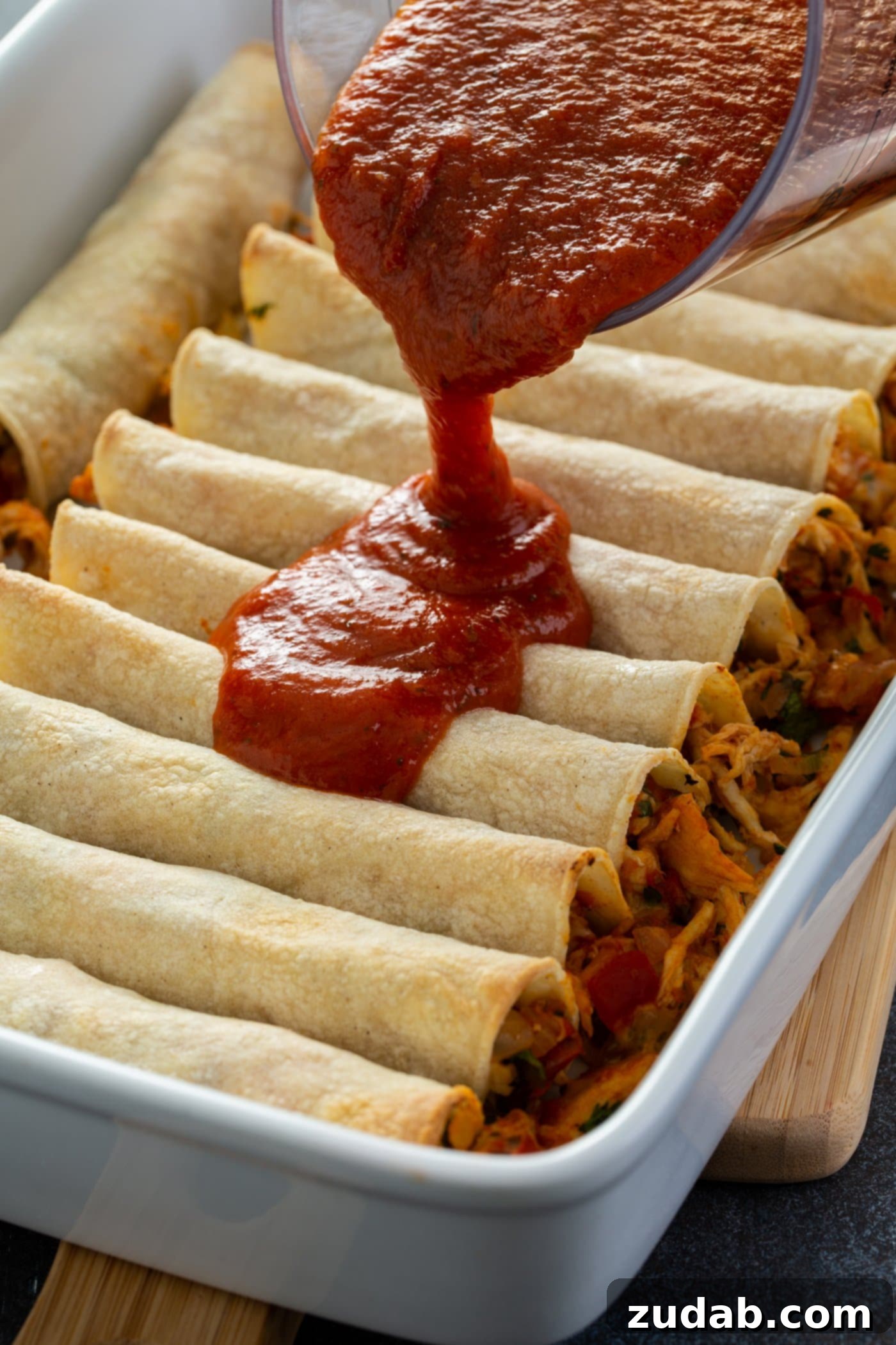 Red Chicken Enchiladas A baking dish filled with chicken enchiladas. A red sauce is being poured over the uncooked enchiladas.