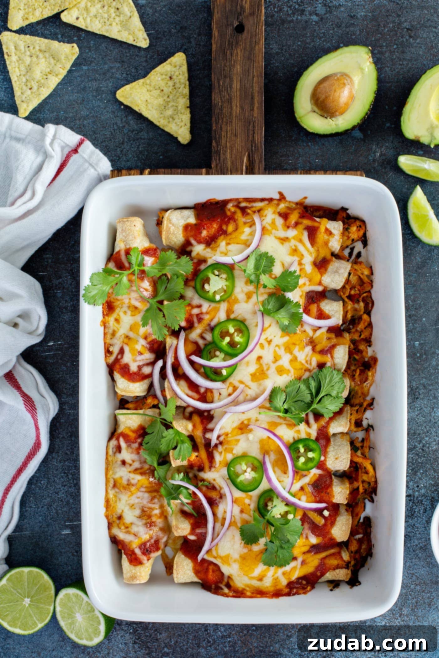Red Chicken Enchiladas A white baking dish filled with baked red chicken enchiladas. They are topped with red sauce, melted cheese, cilantro leaves, slices of red onion and slices of jalapeno. There are lime wedges, halved avocados, and a white with red striped kitchen towel in the background.