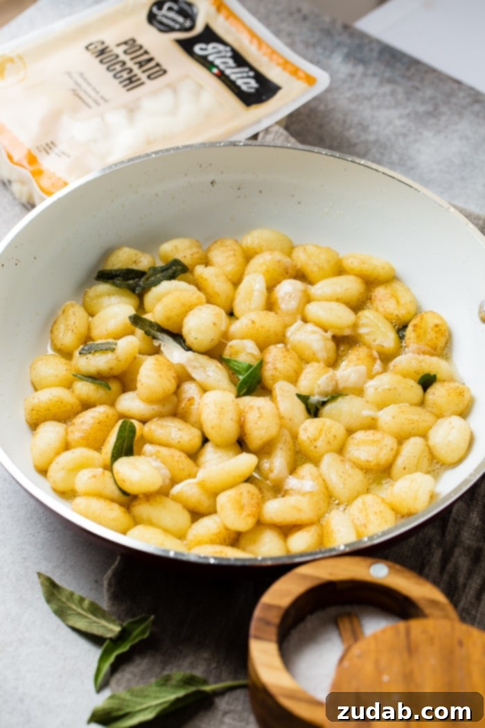 Easy 10-Minute Brown Butter Sage Gnocchi 4 A white skillet with brown butter, gnocchi, and fresh sage to make a yummy 10 minute brown butter sage gnocchi recipe with a package of gnocchi in the background and a container of salt in front.