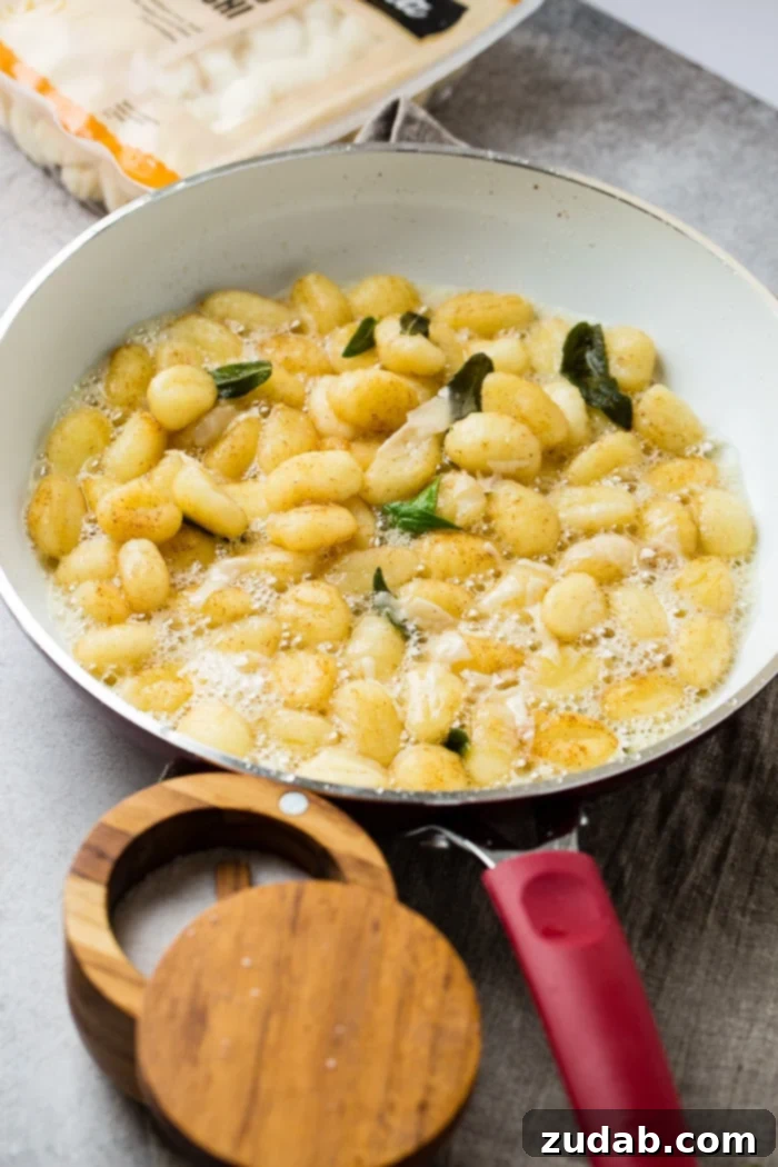 Easy 10-Minute Brown Butter Sage Gnocchi 7 A white skillet with brown butter, gnocchi, and fresh sage to make a yummy 10 minute brown butter sage gnocchi recipe with a package of gnocchi in the background and a container of salt in front.