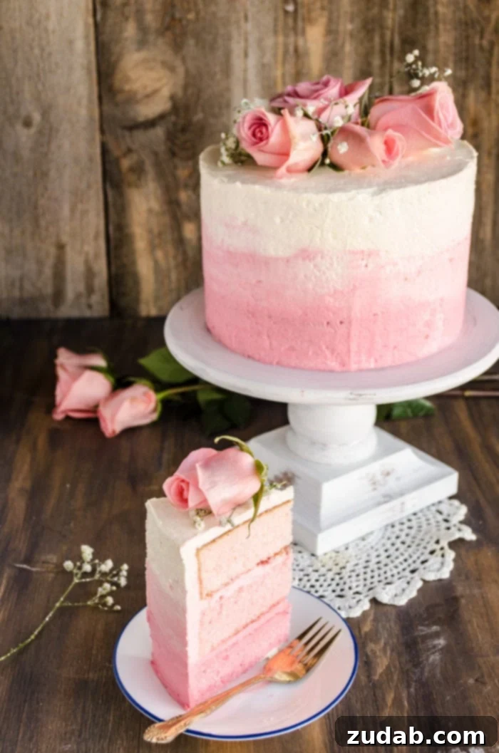 Blushing Strawberry Ombre Cake 2 A stunning three-layer pink ombre cake topped with fresh roses and delicate sprigs of baby's breath, elegantly presented on a raised cake platter. A perfect slice of the ombre cake, showcasing its beautiful gradient of pink layers, rests on a plate beside a fork, inviting a delightful indulgence.