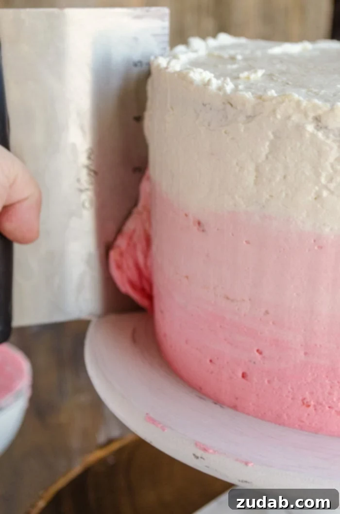 Blushing Strawberry Ombre Cake 12 A three-layer Mother's Day Strawberry cake is topped with three shades of pink frosting. A hand holding a bench scraper is smoothing the sides of the cake, blending the ombre colors.