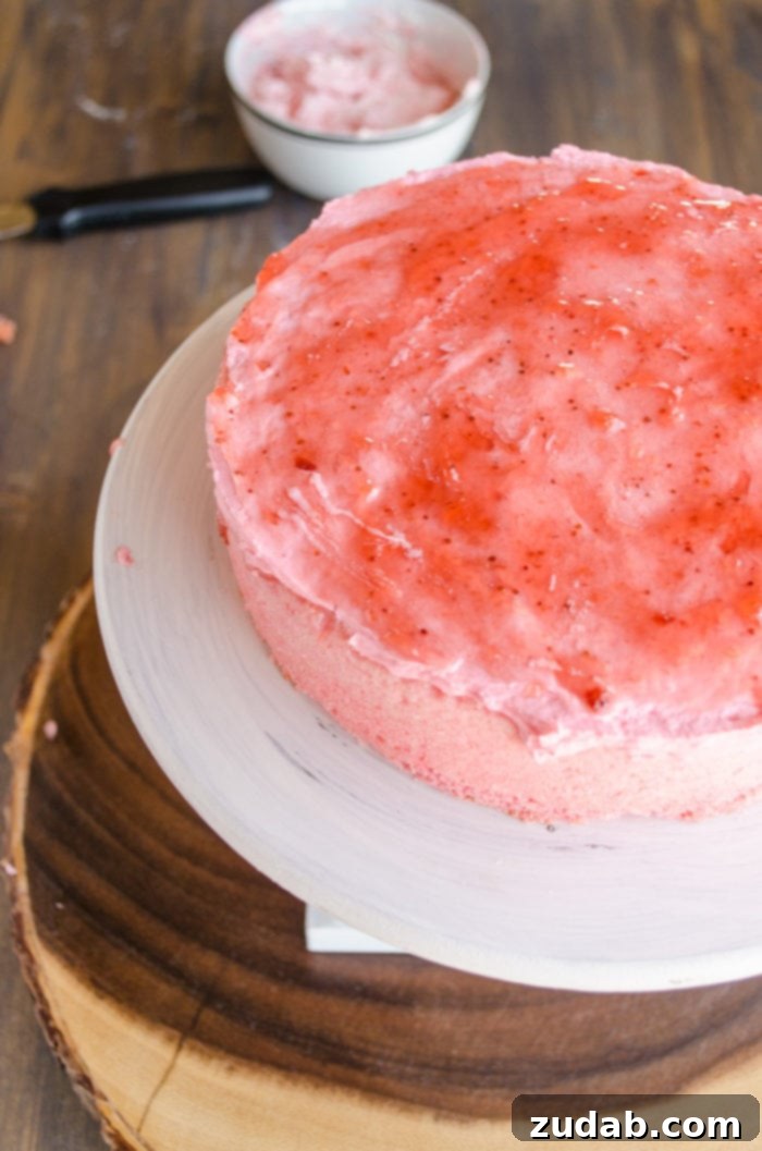 Blushing Strawberry Ombre Cake 10 The deepest pink layer of the cake is positioned on a raised cake stand, with a corresponding pink frosting generously spread on top, crowned with a layer of glistening strawberry jam. In the background, a bowl of pink frosting awaits its turn.