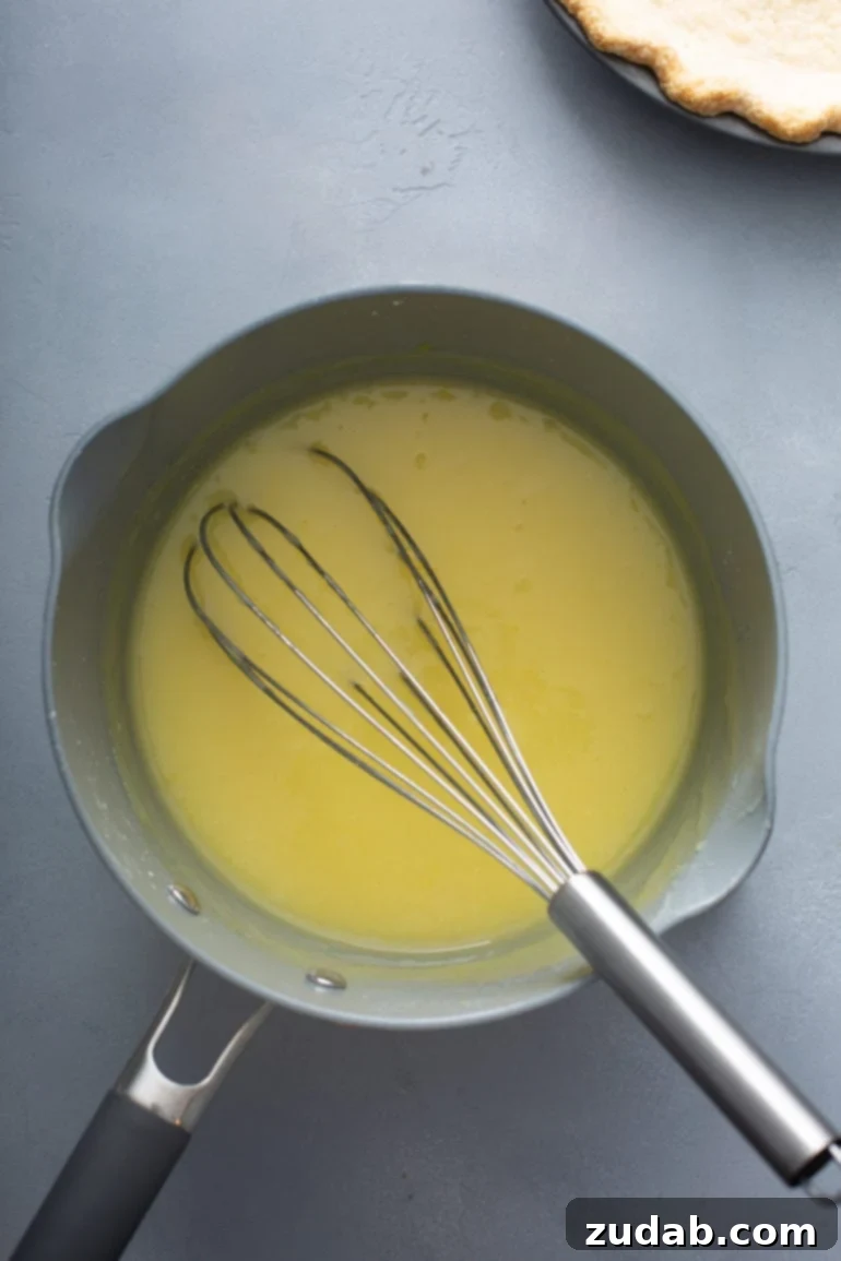 Mom's Secret Lemon Meringue Pie 7 A vibrant lemon pie filling in a saucepan, being stirred with a whisk, highlighting the final stages of cooking and butter incorporation.