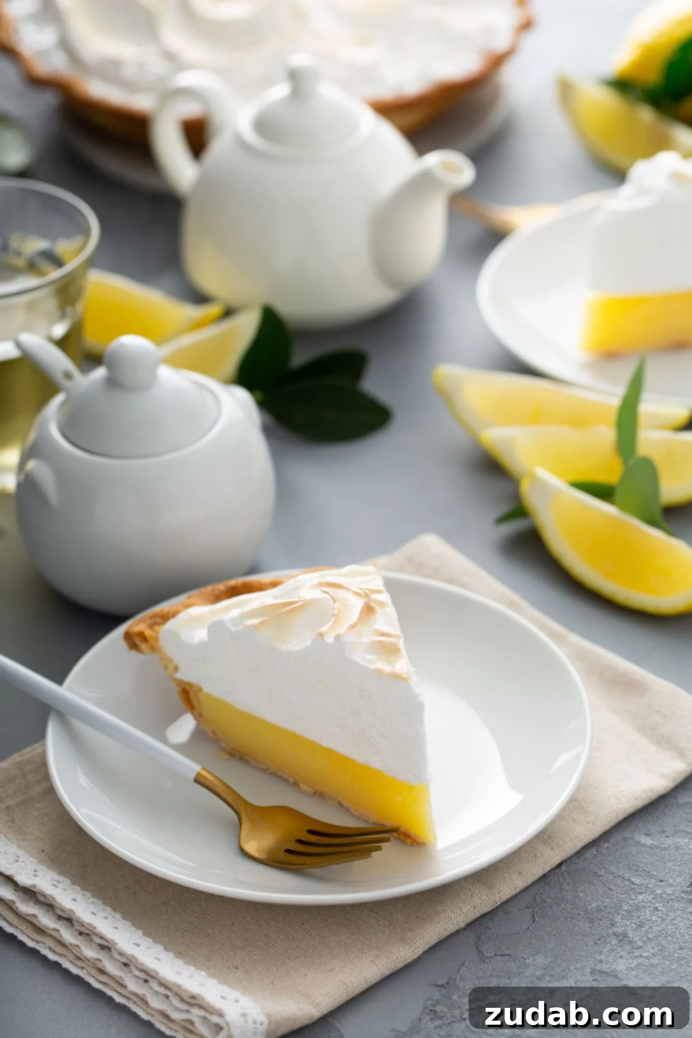 A Delicious Slice of Lemon Meringue Pie A mouth-watering piece of lemon meringue pie on a pristine white dessert plate. A fork is placed invitingly next to the pie, with fresh lemon wedges, a sugar bowl, and a cream pitcher adding charm to the background.