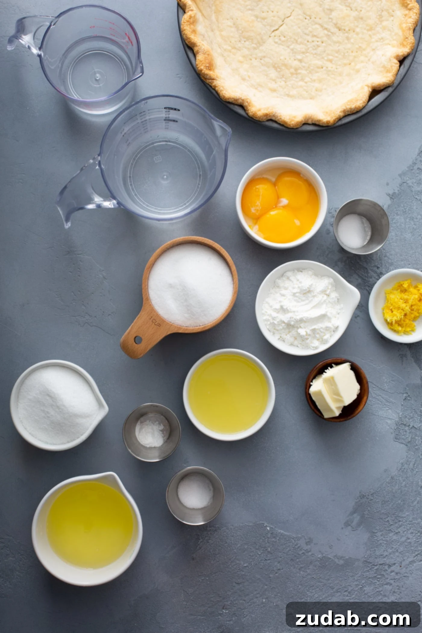 Fresh Ingredients for Lemon Meringue Pie A collection of measuring cups and small bowls filled with ingredients for lemon meringue pie. Fresh lemons, eggs, sugar, butter, and water are neatly arranged, showcasing the wholesome components of the recipe.