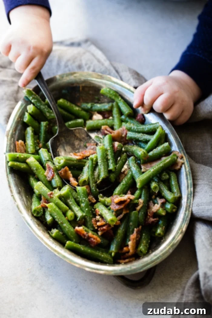 Quick Bacon Green Beans 6 A toddler boy's hands reaching across a grey table, ready to use a vintage silver serving spoon to scoop chopped green beans with crispy bacon from a silver serving bowl.