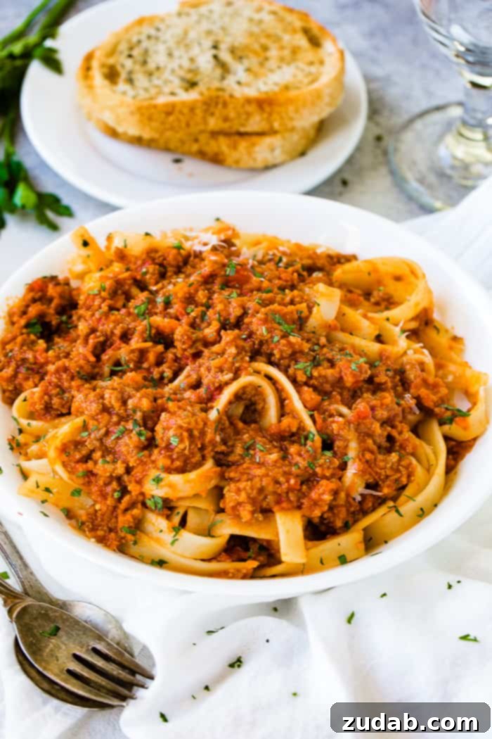 Mastering Authentic Italian Bolognese 7 A close-up of a serving of fettuccine pasta coated in authentic Italian Bolognese sauce, garnished with fresh Parmesan cheese, with a blurred background of garlic bread.
