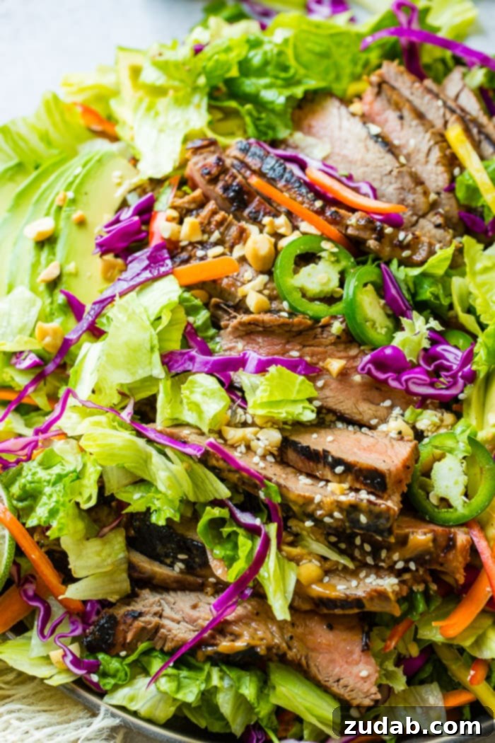 Grilled Flank Steak with Zesty Thai Peanut Salad 6 A grey plate filled with mixed greens, fresh vegetables, and tender flank steak marinated in a rich, creamy nut-based Thai peanut sauce, drizzled with a zesty ginger dressing and garnished with sesame seeds.