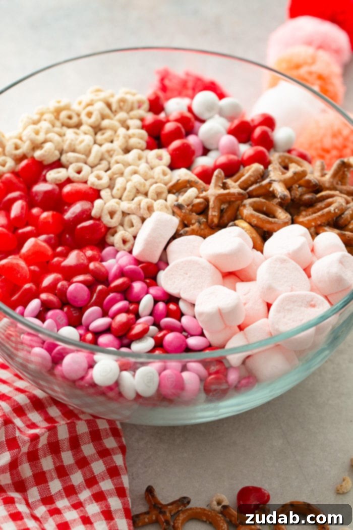 5-Minute Valentine's Treat Mix 3 A close-up of a large glass bowl filled with a colorful array of Frosted Cheerios, Valentine's caramel M&Ms, X and O shaped pretzels, pink heart marshmallows, Valentine's colored plain M&Ms, and heart shaped Starburst jelly beans, creating an inviting Valentine's Day snack mix.