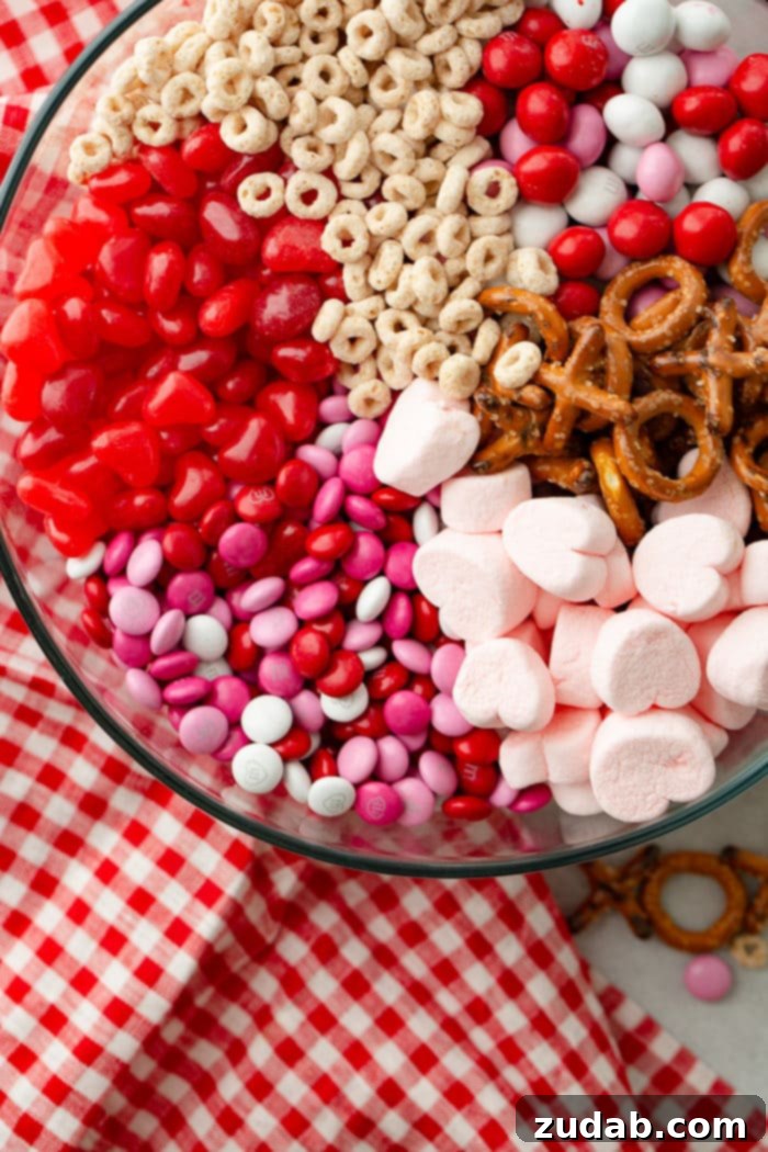 5-Minute Valentine's Treat Mix 6 A festive Valentine's snack mix presented in a large glass bowl, featuring frosted Cheerios, Valentine's caramel M&Ms, X and O shaped pretzels, pink heart marshmallows, Valentine's colored plain M&Ms, and heart-shaped Starburst jelly beans, artfully arranged beside a charming red and white checkered napkin.