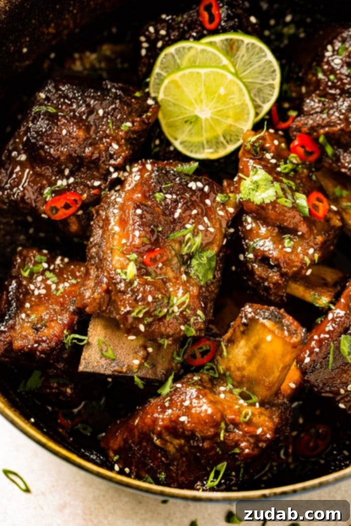 Tender Oven Braised Korean Beef Short Ribs 3 A photo of several braised short ribs still in the dutch oven garnished with sesame seeds, red chile peppers, cilantro, green onions, and lime slices.