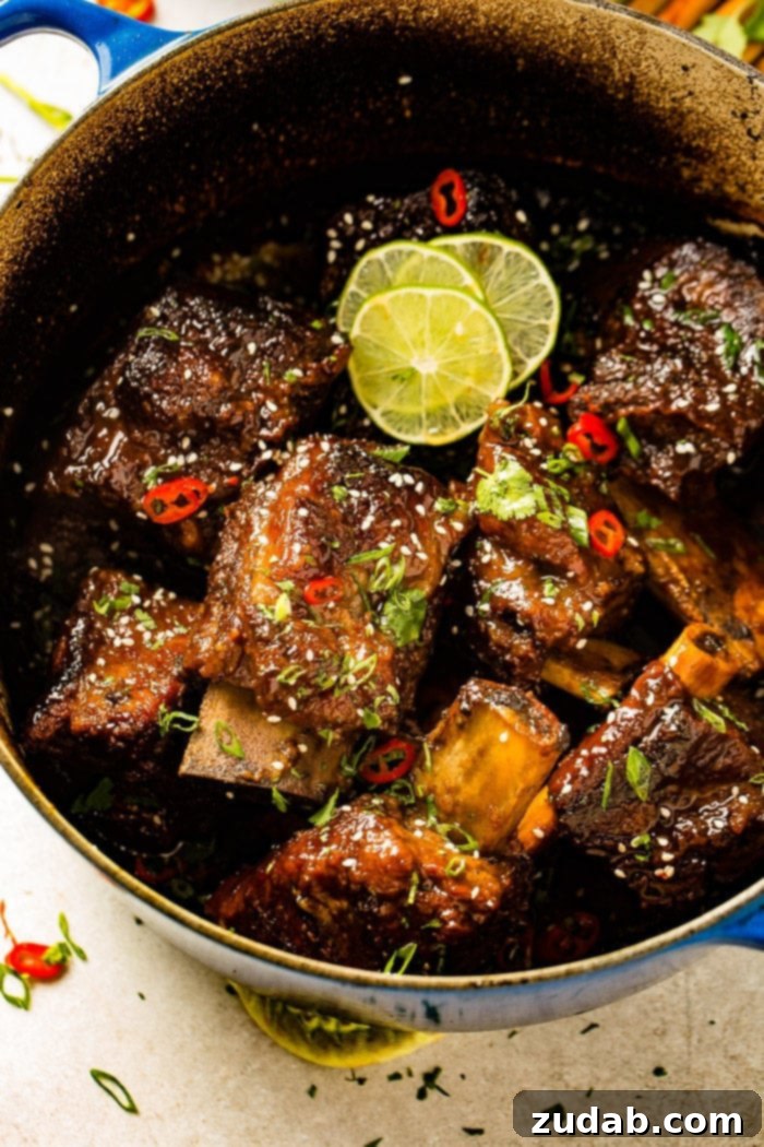Tender Oven Braised Korean Beef Short Ribs 7 A photo of several braised short ribs still in the dutch oven garnished with sesame seeds, red chile peppers, cilantro, green onions, and lime slices.