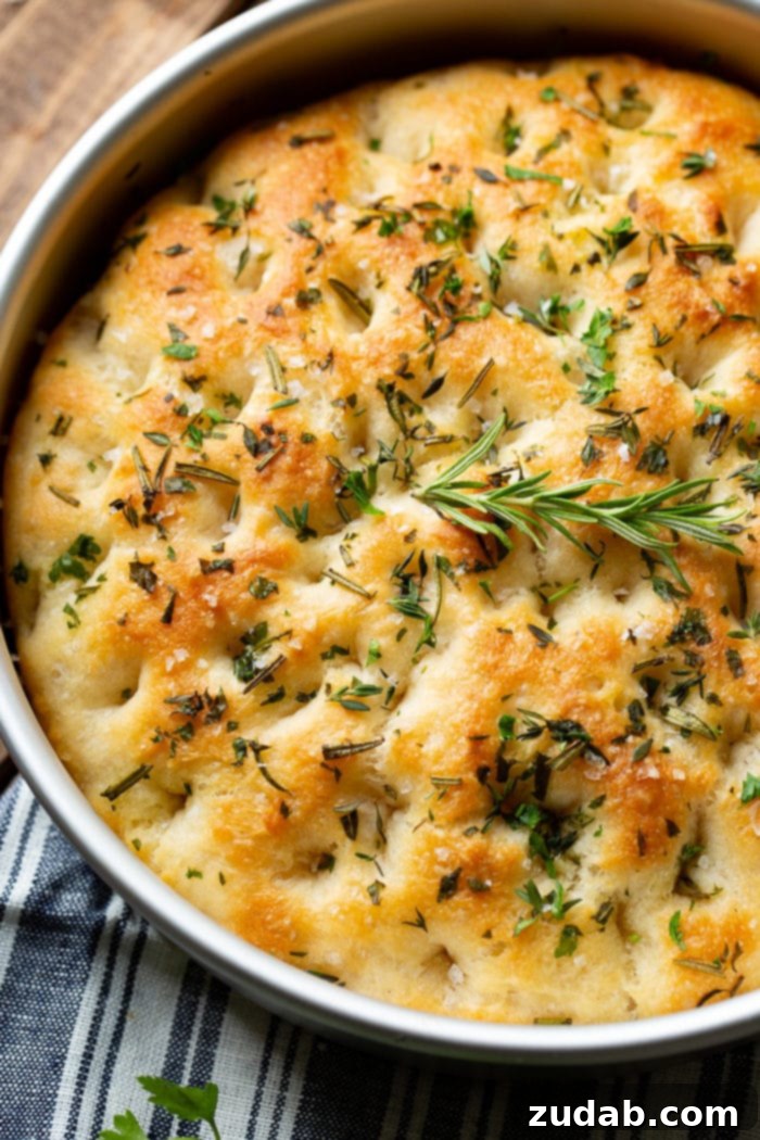 Effortless Aromatic Focaccia Bread 5 A round focaccia bread loaf, golden and rustic, topped with a mix of fresh rosemary, thyme, and flaky sea salt. It's nestled in a silver baking pan, ready to be served or cooled.