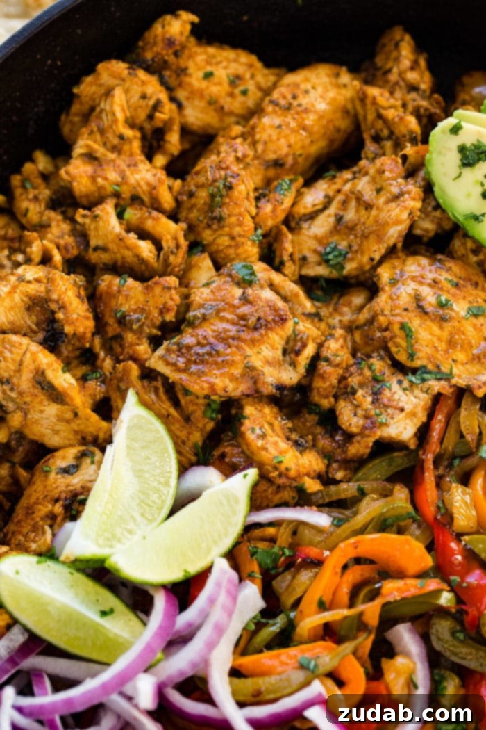 Family-Friendly One-Skillet Chicken Fajitas 7 A photo of a cast iron skillet full of chicken fajita filling - cooked chicken, sauteed peppers and onions, lime wedges, sliced purple onion, and sliced avocados.