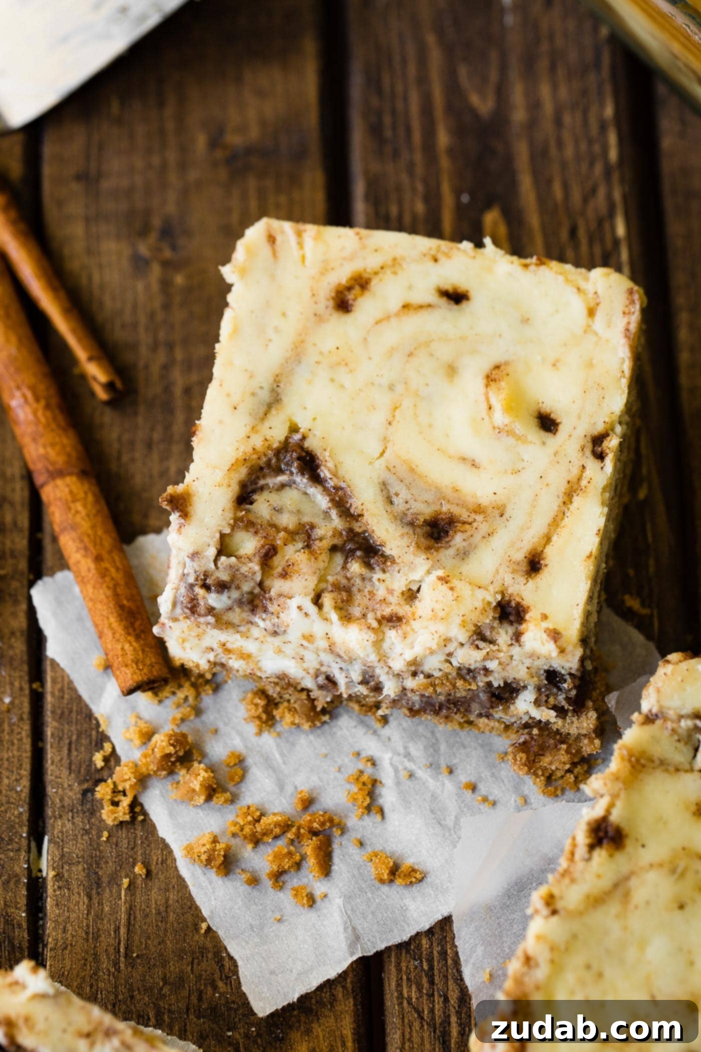 Blender Cinnamon Roll Cheesecake Bars A photo of a cinnamon roll cheesecake bar sitting on a wooden surface with cinnamon sticks sitting next to it, highlighting the textures.
