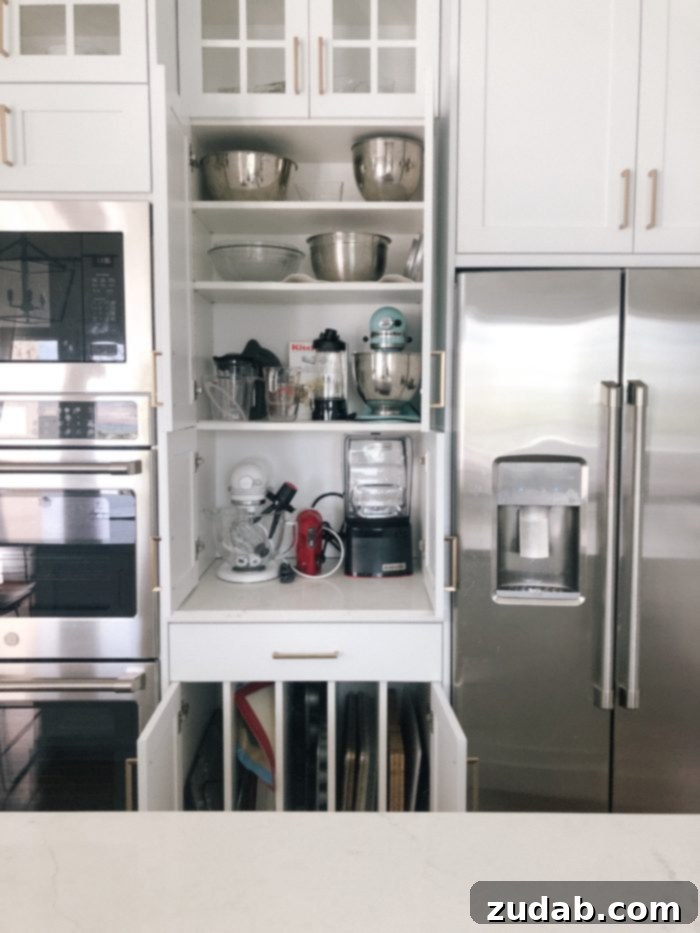 Detailed view of an all-white kitchen's long island, white counters, glass-door cabinets, black pendant lights, pantry, and floating wood shelves
