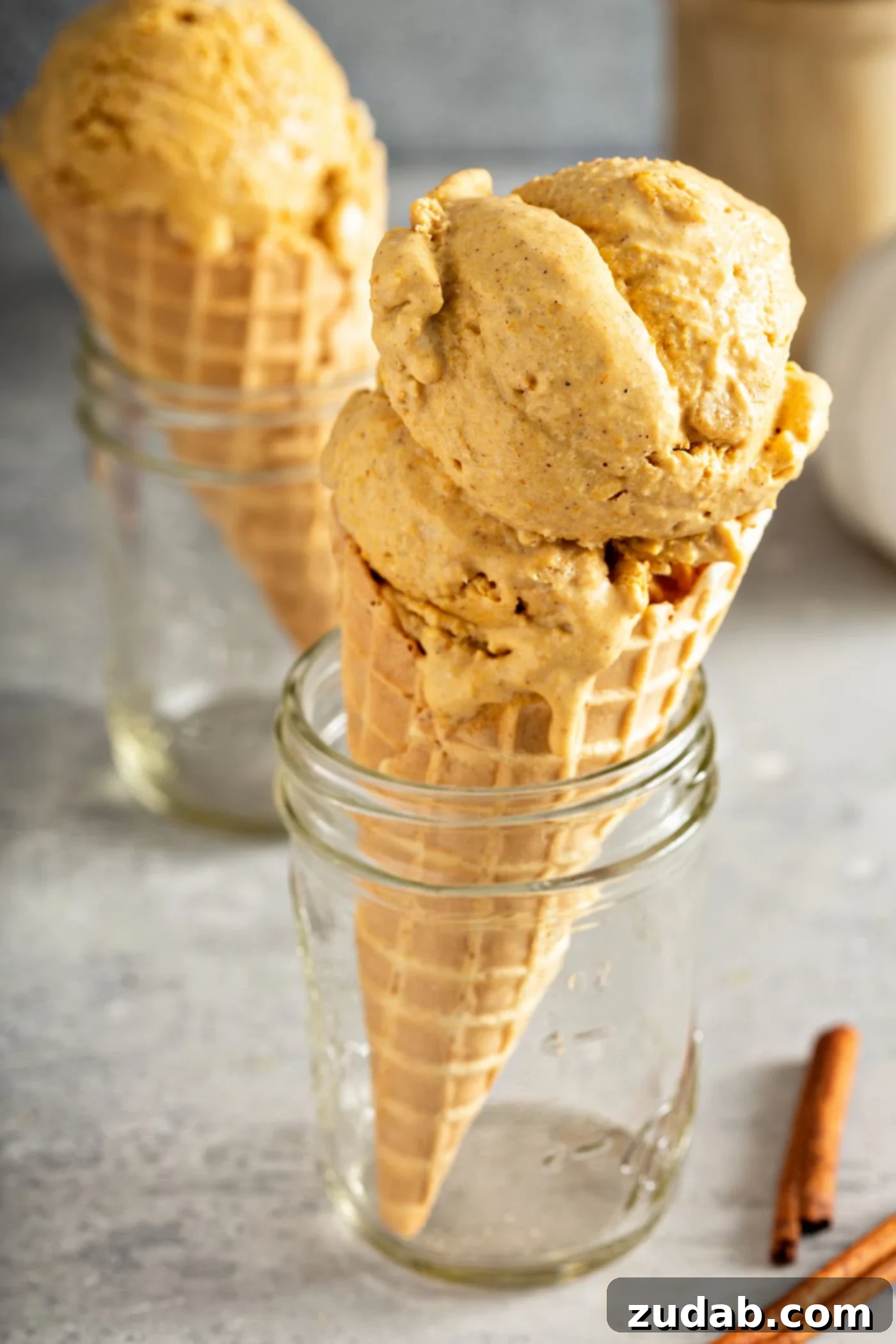 The Creamiest Pumpkin Ice Cream A sugar cone with two scoops of pumpkin ice cream. The cone is propped in a jar. There is another ice cream cone in the background and a few cinnamon sticks next to the ice cream cone.