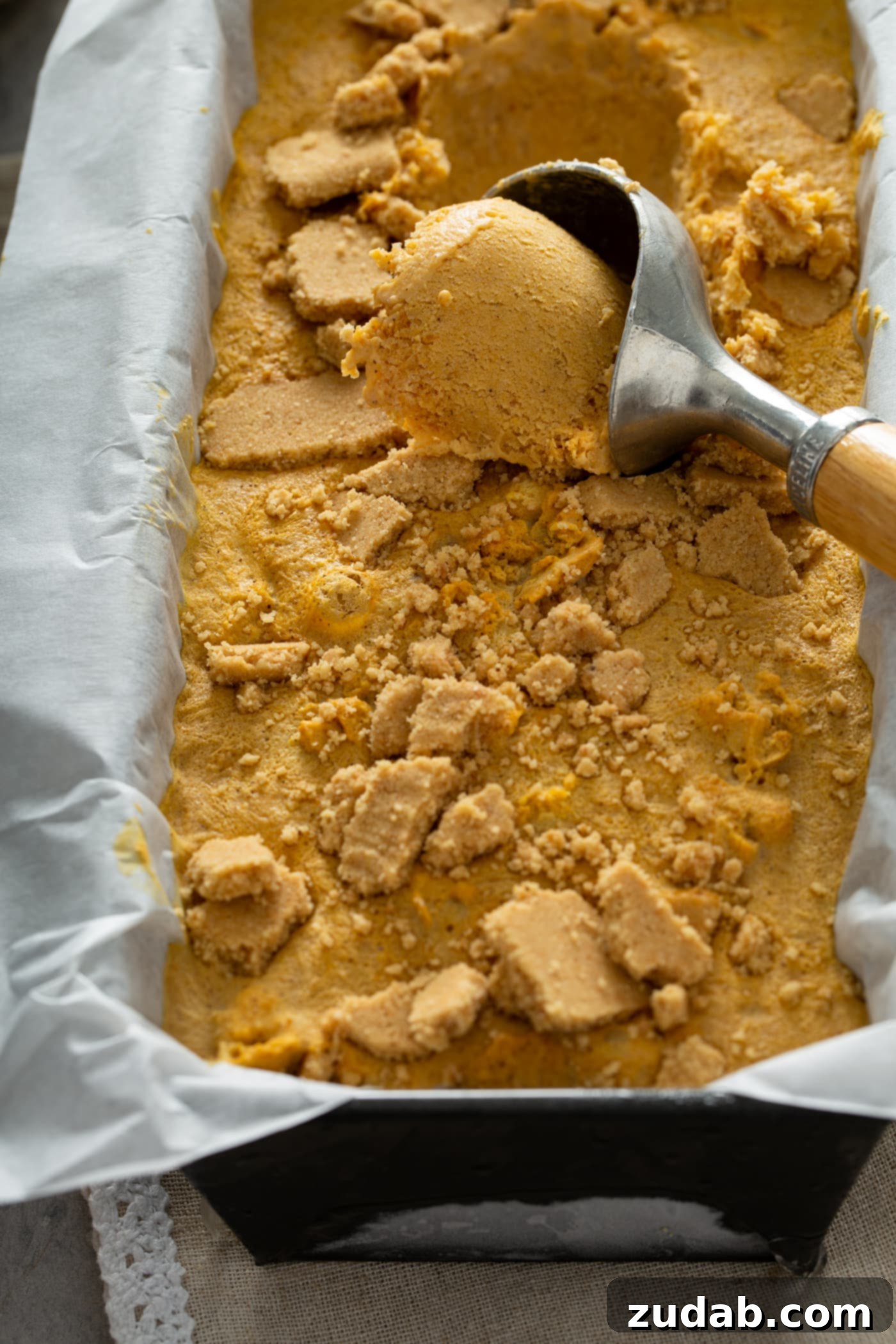 The Creamiest Pumpkin Ice Cream A loaf pan lined with parchment paper and filled with pumpkin ice cream. There is an ice cream scoop with a scoop of ice cream in it.