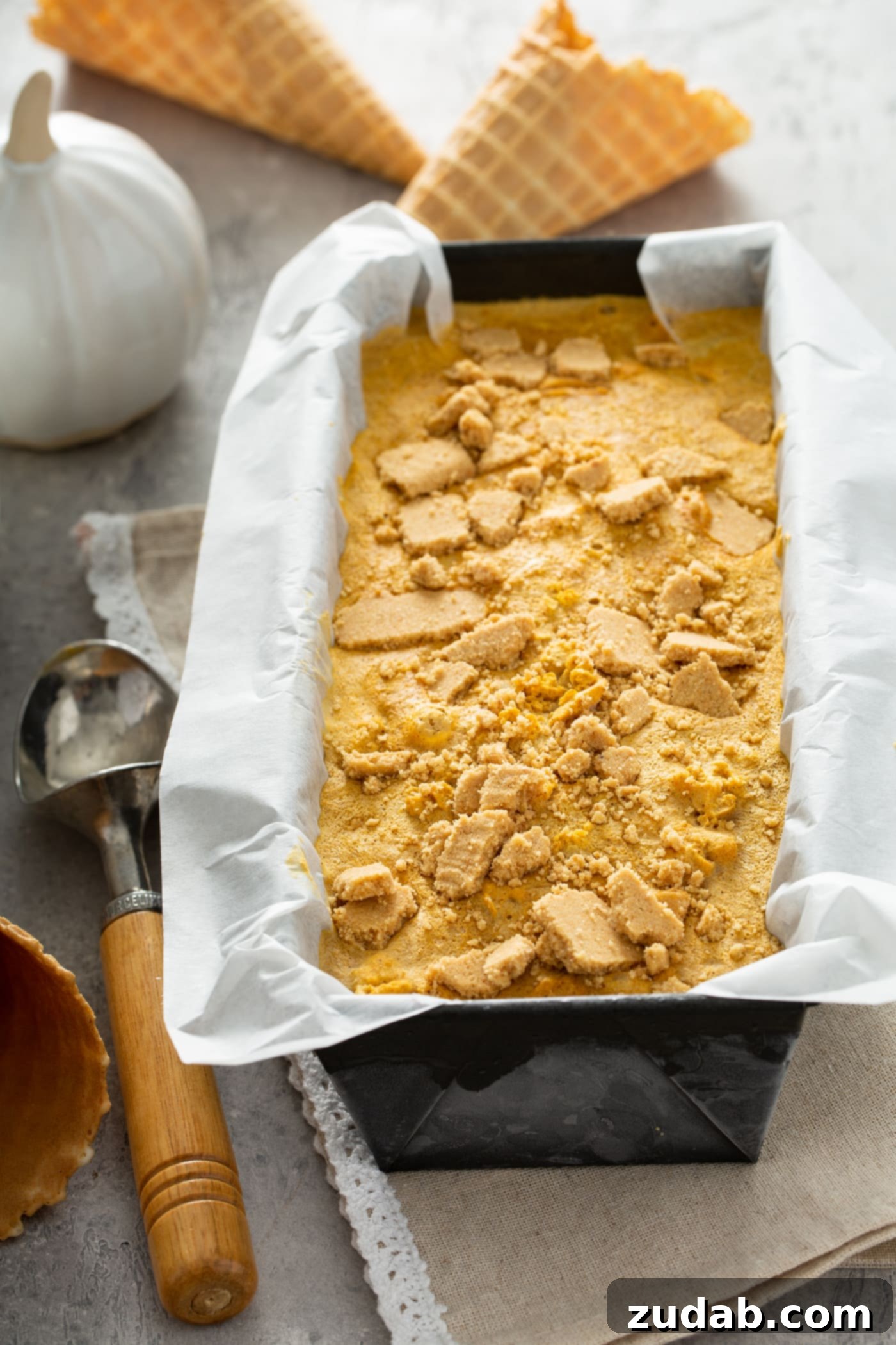 The Creamiest Pumpkin Ice Cream A loaf pan lined with parchment paper and filled with pumpkin ice cream with graham cracker crust sprinkled on top. There is a spoon next to the pan of ice cream and a tiny white pumpkin in the background.