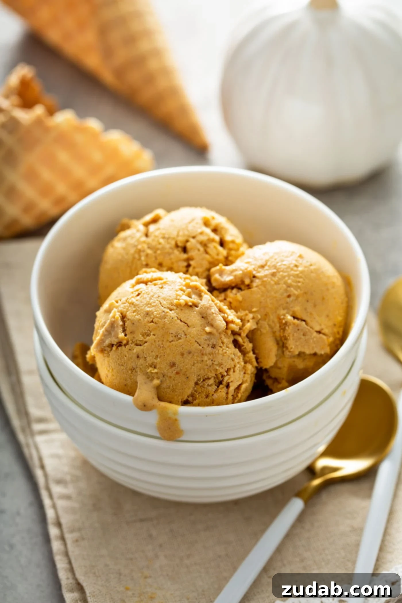 The Creamiest Pumpkin Ice Cream A white dessert bowl with three scoops of pumpkin ice cream. There is a spoon next to the bowl which is sitting on a napkin. A sugar cone and a tiny white pumpkin are in the background.