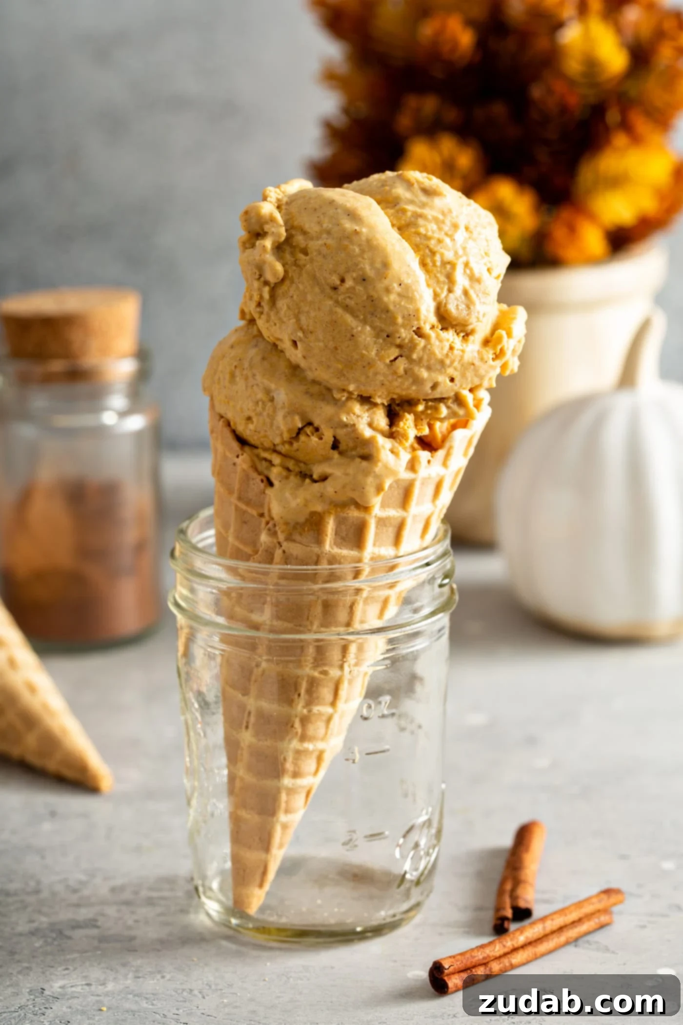 The Creamiest Pumpkin Ice Cream A sugar ice cream cone with two scoops of pumpkin ice cream. The cone is sitting in a glass jar and there is another cone laying on the table in the background with a container of cinnamon, a small white pumpkin and cinnamon sticks.