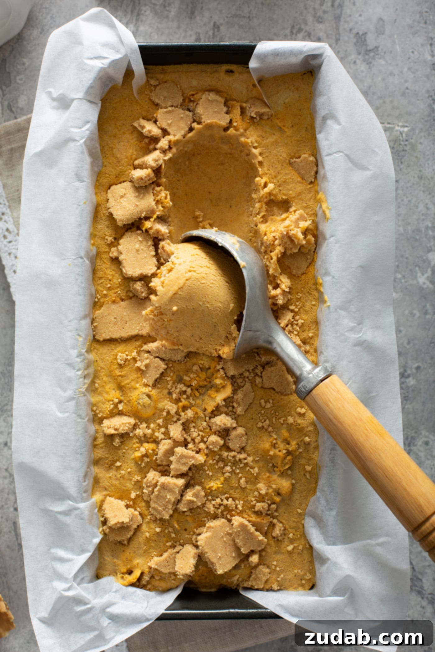 The Creamiest Pumpkin Ice Cream A scoop of ice cream being removed from a loaf pan lined with parchment paper and filled with pumpkin ice cream.
