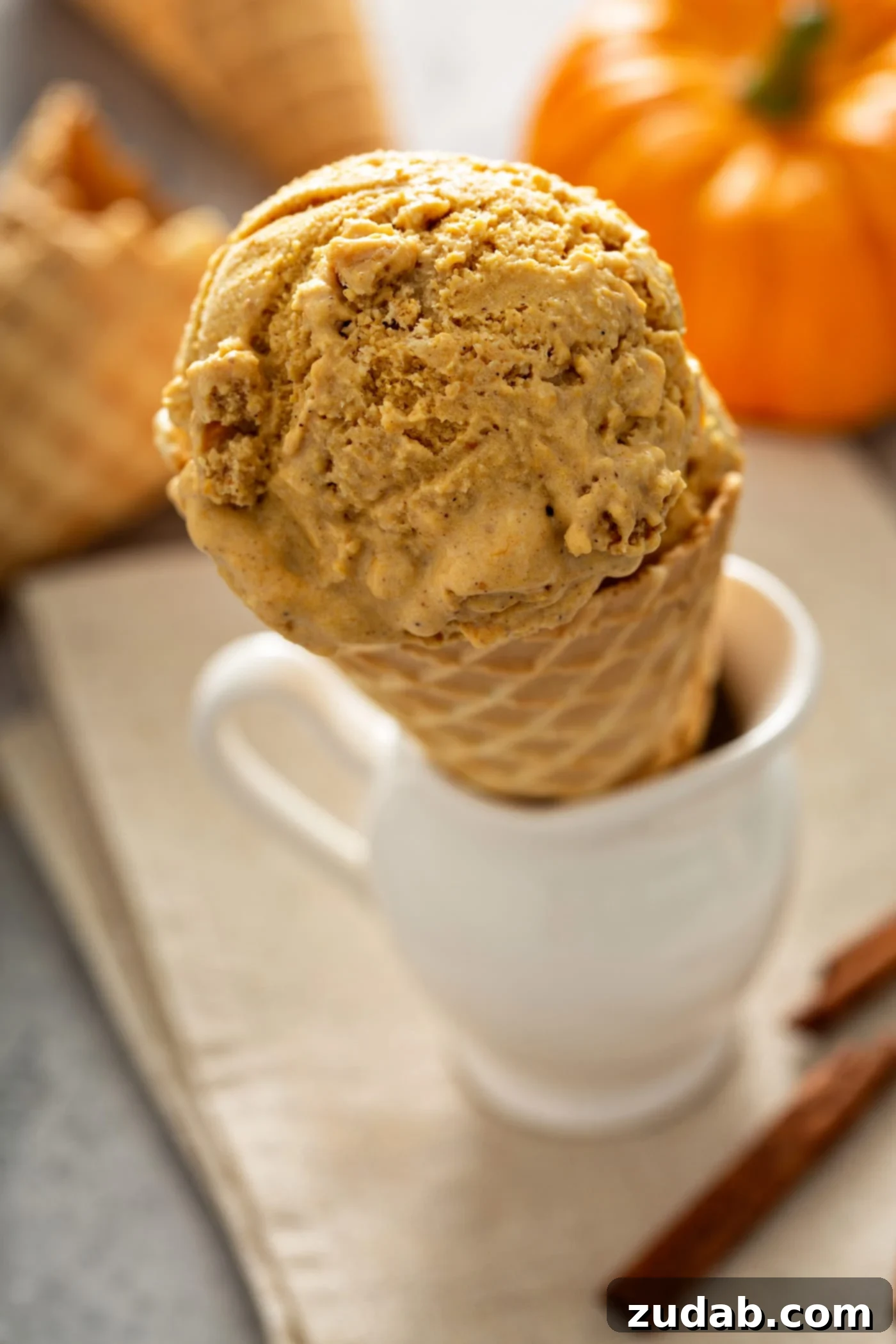 The Creamiest Pumpkin Ice Cream A scoop of pumpkin ice cream in a sugar cone that is standing in a white cup. There are cinnamon sticks and other cones in the background.