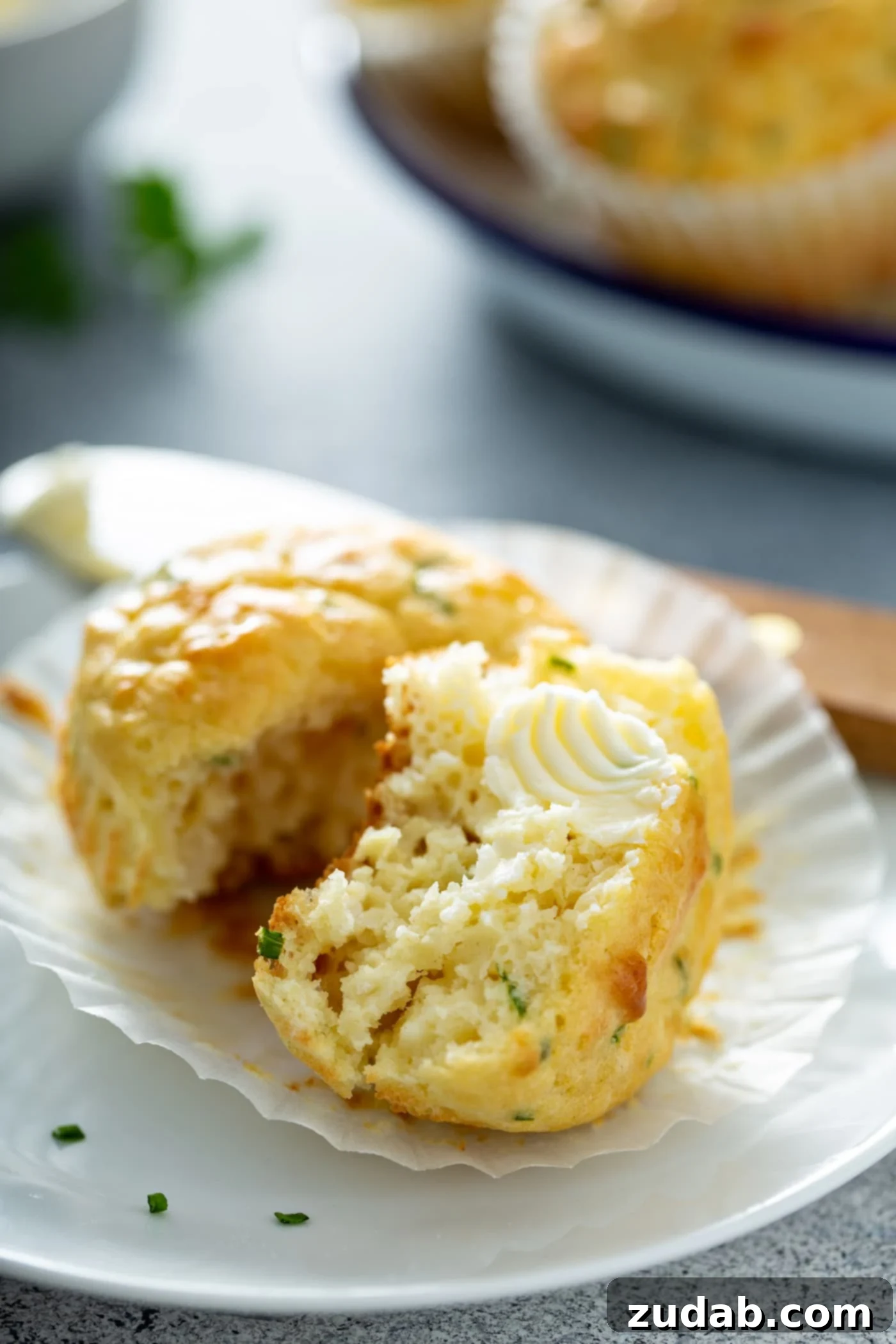 Quick and Easy Feta Muffins A muffin with the paper muffin liner peeled back. The muffin is broken in half and you can see the herbs inside. There is a butter knife and a plate of muffins in the background.