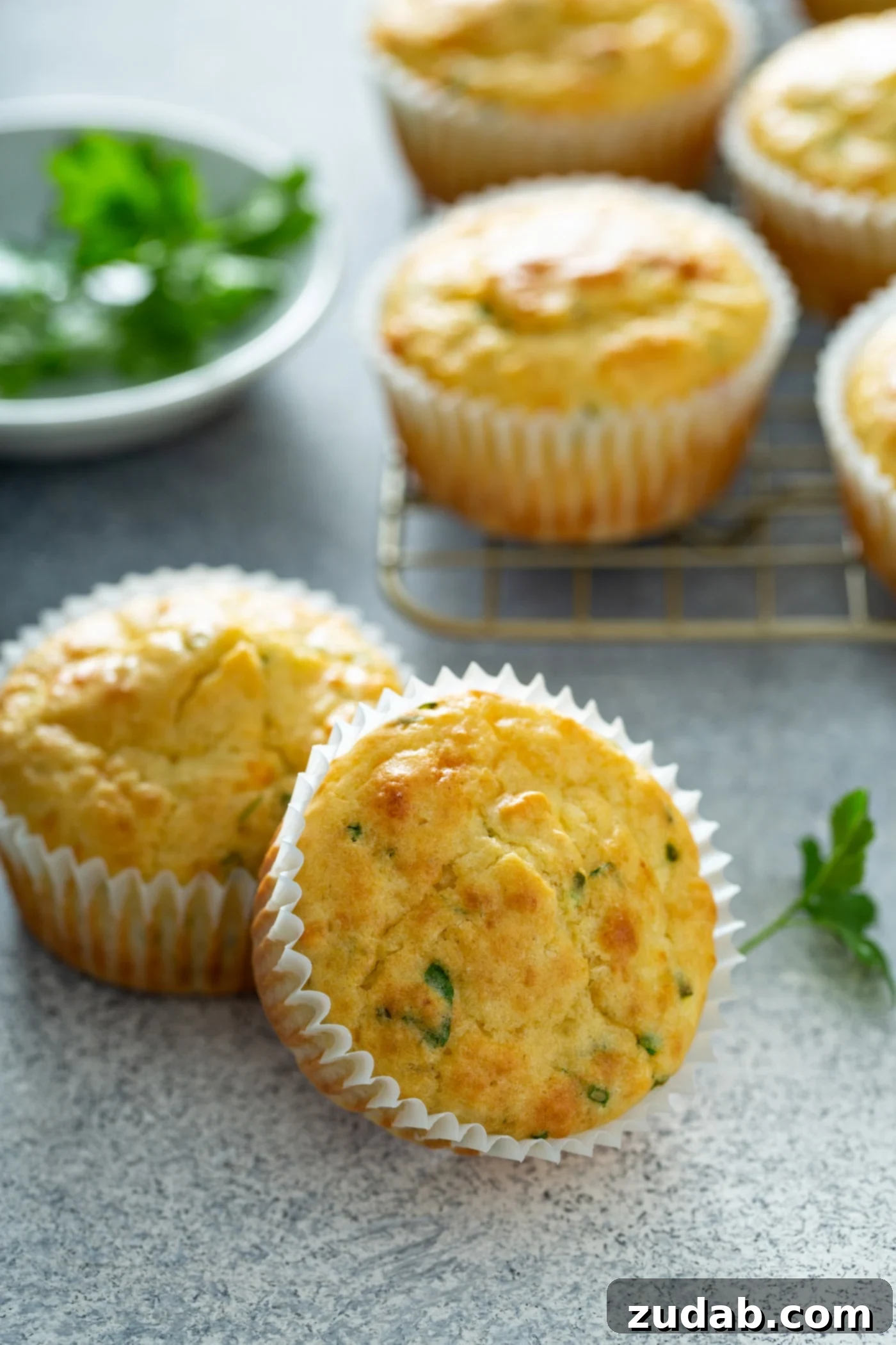 Quick and Easy Feta Muffins Four baked feta muffins in white paper liners. There is a bowl of chopped parsley and chives in the background.