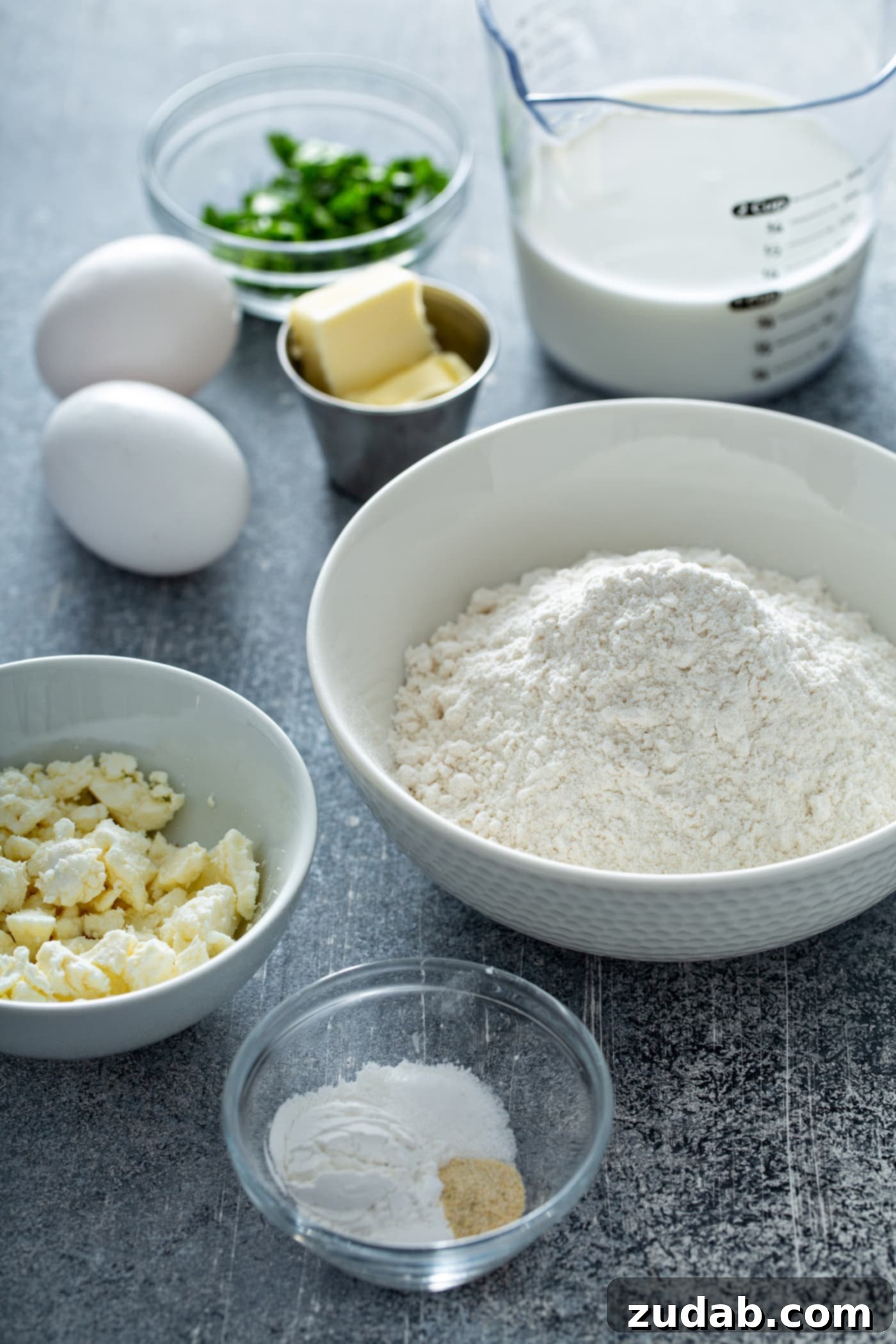 Quick and Easy Feta Muffins A table top with containers of Flour, feta cheese, butter, salt, green herbs and two white eggs.