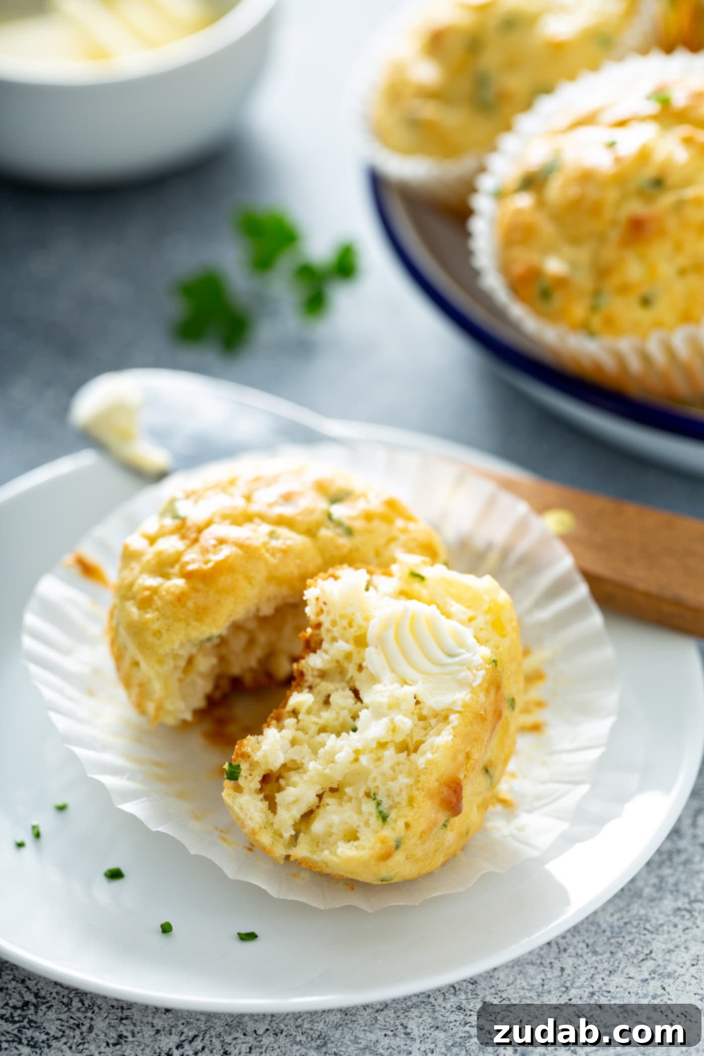 Quick and Easy Feta Muffins A muffin on a plate. The paper muffin liner is peeled back and the muffin is broken in half showing the herbs inside. A pat of butter is on one half of the muffin. There is a butter knife resting on the plate that the muffin is on. There is a plate of muffins in the background.