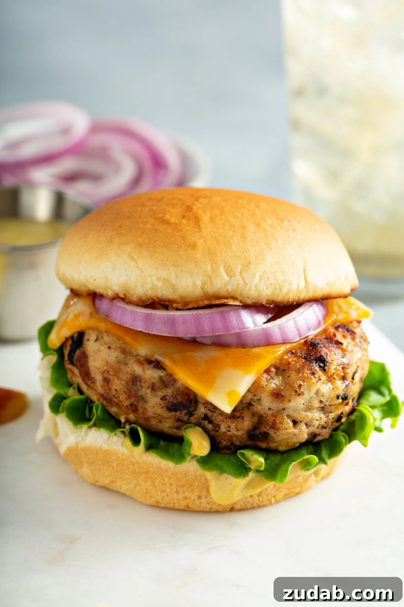 Maple Mustard Apple Chicken Burgers A perfectly grilled maple mustard apple chicken burger on a toasted brioche bun, layered with fresh lettuce, melted Monterey cheese, and thinly sliced red onions. In the background, a plate of vibrant sliced red onions adds a pop of color, highlighting the fresh ingredients.