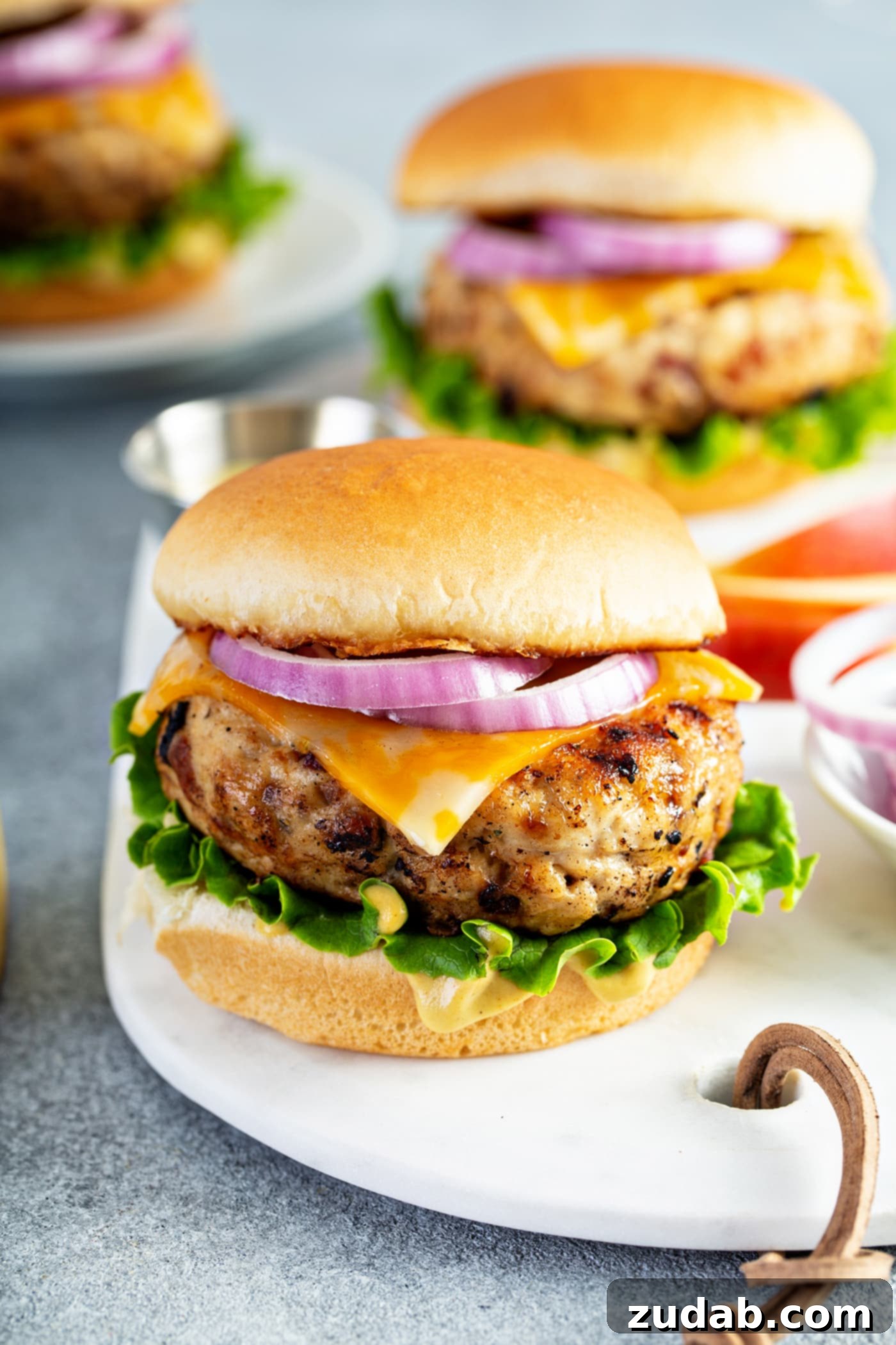 Maple Mustard Apple Chicken Burgers Three perfectly plated dinner servings of maple mustard apple chicken burgers, each nestled in a bun with crisp lettuce, melted cheese, and vibrant red onions. The inviting presentation makes them ready to enjoy.