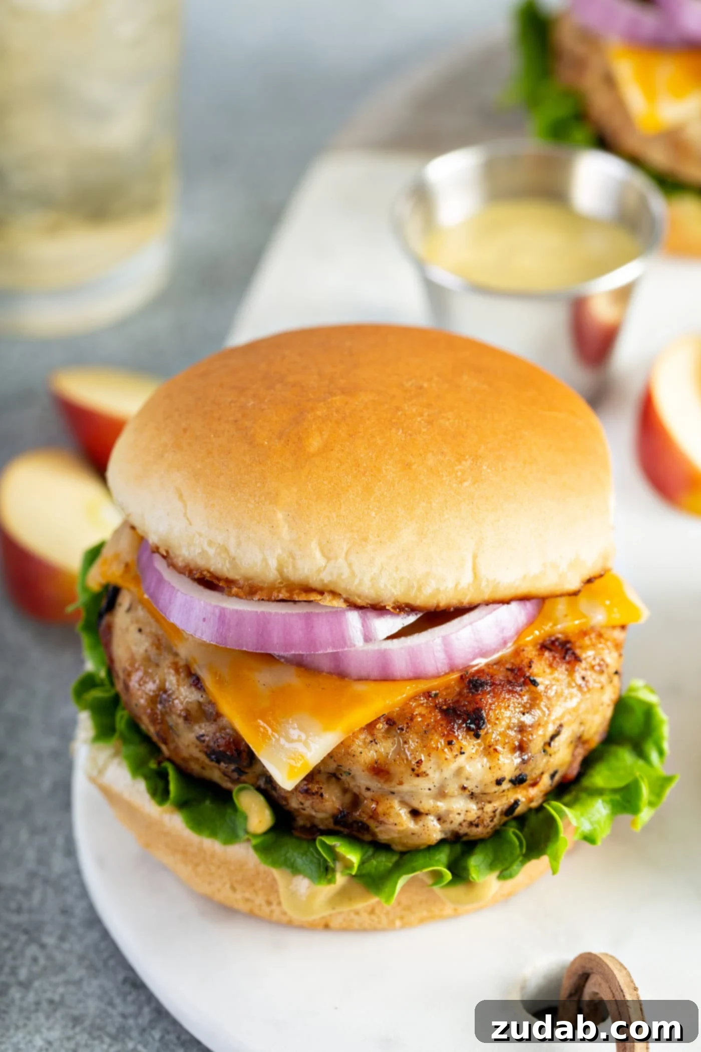 Maple Mustard Apple Chicken Burger A mouth-watering maple mustard apple chicken burger, impeccably presented on a bun with fresh lettuce, melted cheese, and thinly sliced red onion. In the background, vibrant apple slices and a refreshing glass of apple juice complete the inviting scene, emphasizing the apple-infused flavor of the burger.