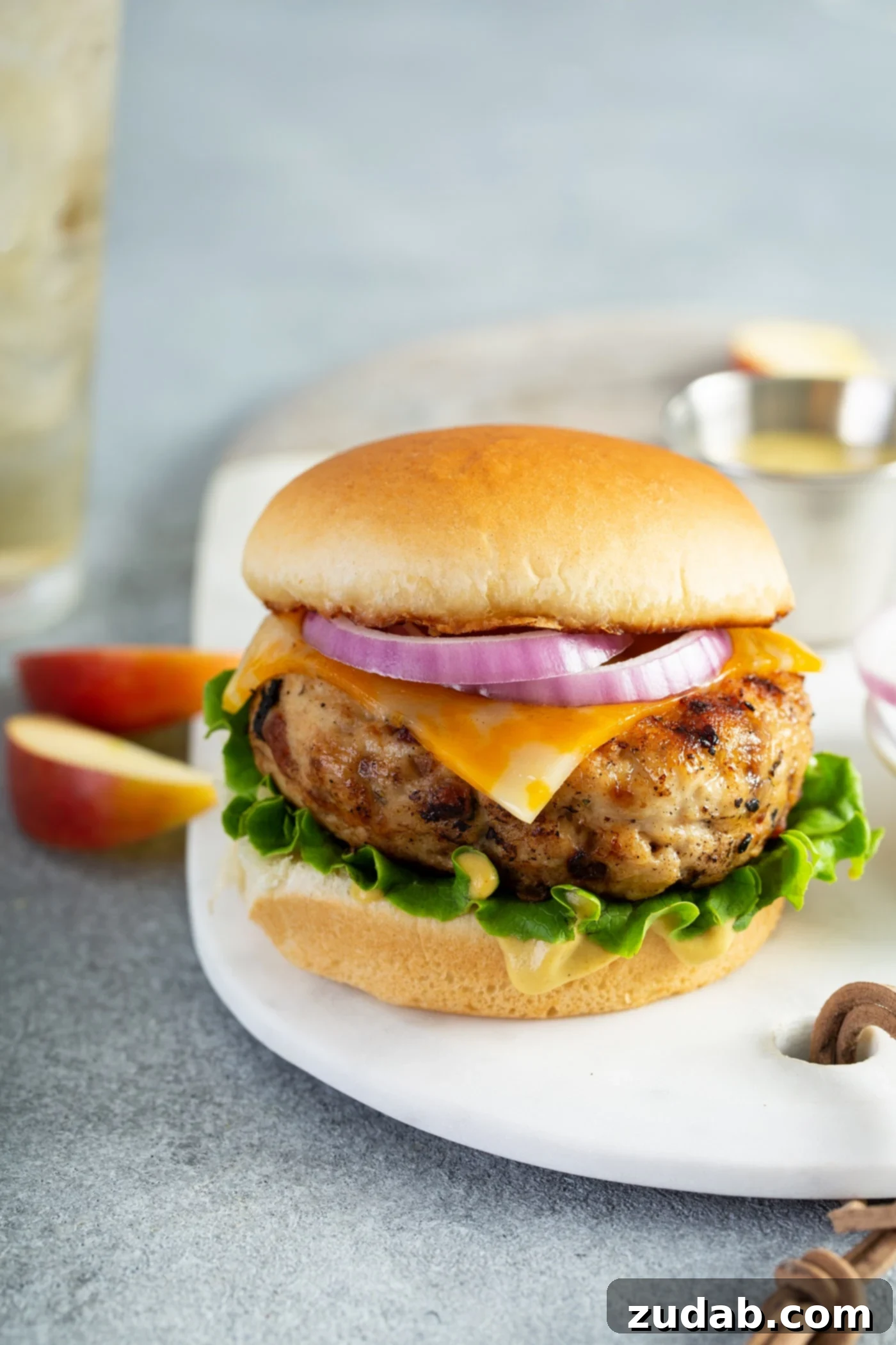 Maple Mustard Apple Chicken Burgers A single, perfectly plated chicken burger on a pristine white plate. The burger is beautifully presented on a toasted bun, generously layered with crisp lettuce, melted cheese, and a vibrant slice of red onion. Two fresh apple slices sit beside the plate, hinting at the unique, sweet and savory flavor profile of the burger.