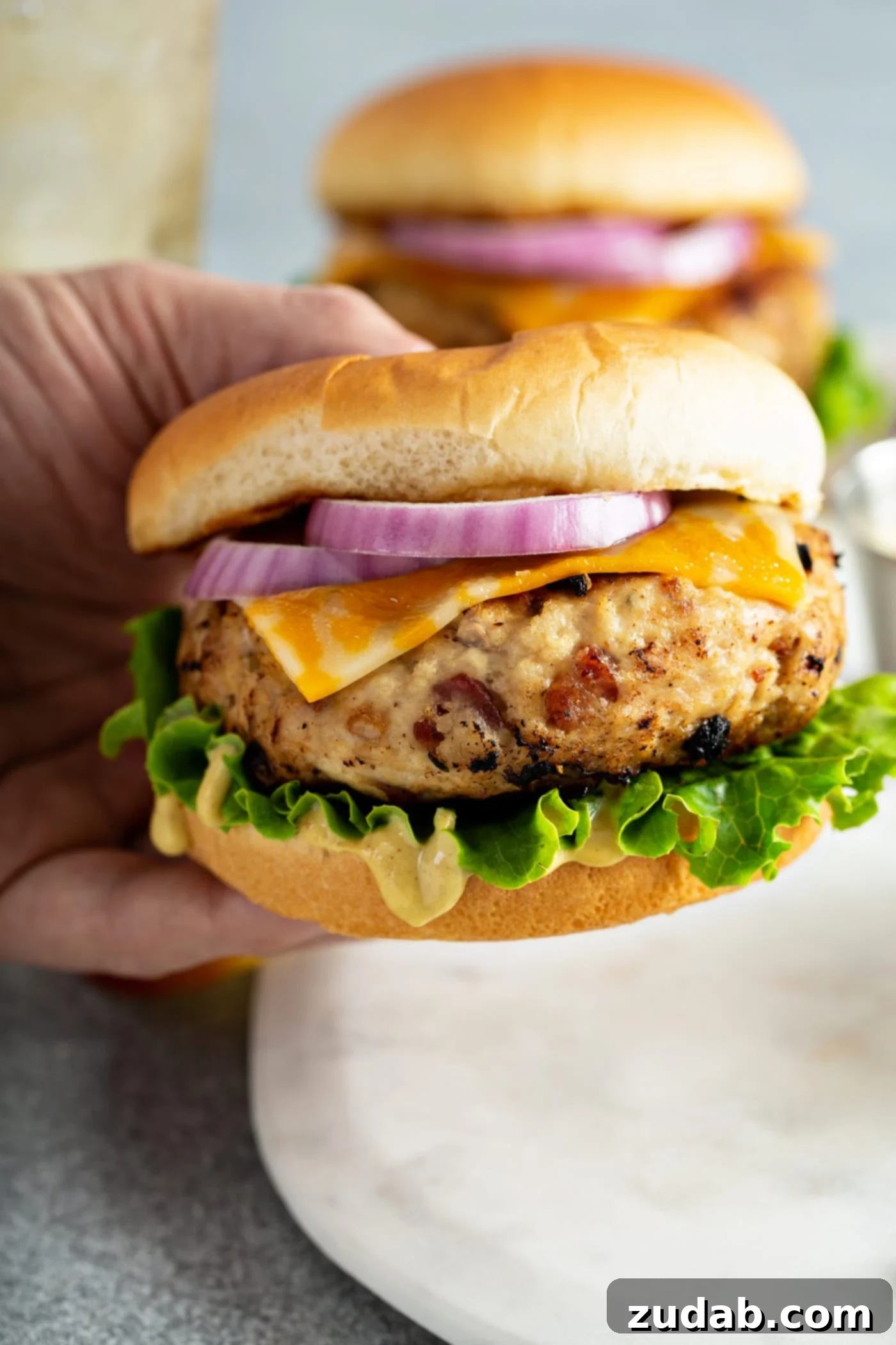 Maple Mustard Apple Chicken Burgers A hand gently holds a delectable chicken burger, showcasing its inviting layers. Nestled within a soft bun, you can see crisp lettuce, melted cheese, and vibrant sliced red onion, all complementing the savory patty. The focus is on the burger's appetizing appeal, promising a delicious bite.