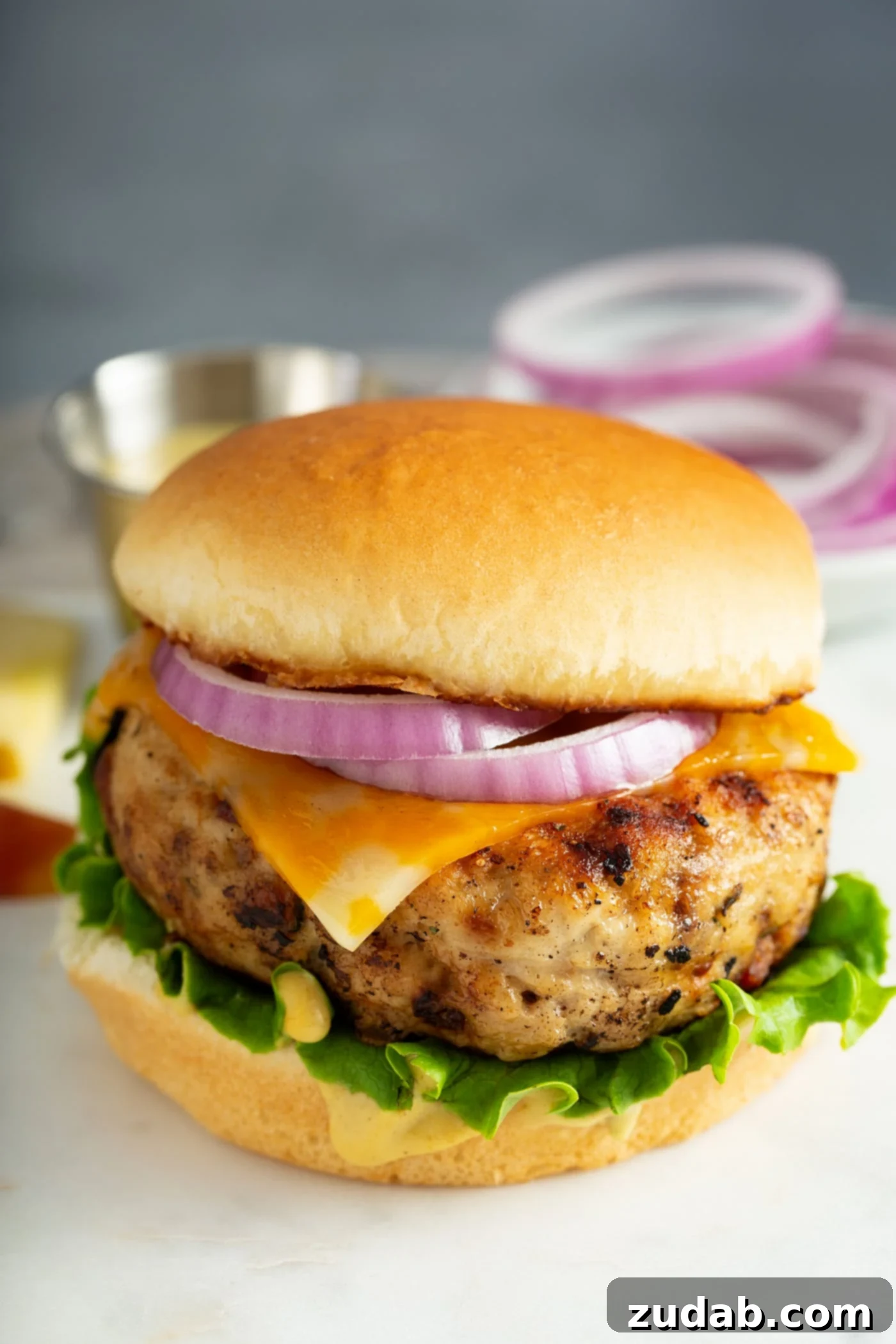 Maple Mustard Apple Chicken Burgers A tantalizing grilled chicken burger, beautifully presented on a soft bun and adorned with crisp lettuce, rich melted cheese, and vibrant sliced red onion. Another mouth-watering chicken burger is visible in the background, hinting at a feast.