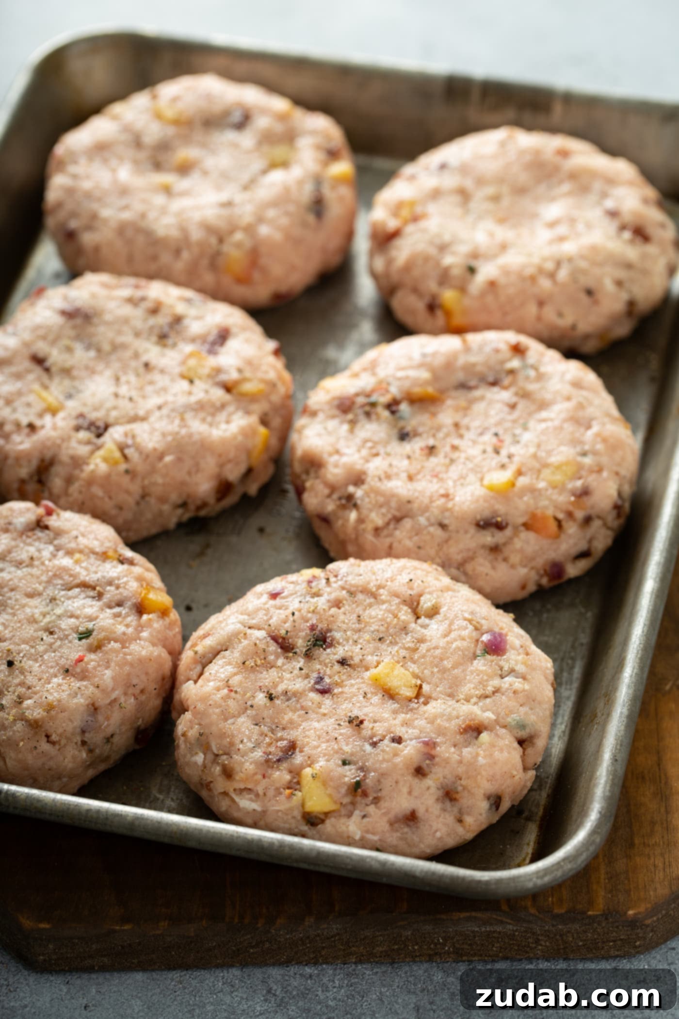 Maple Mustard Apple Chicken Burgers Six perfectly formed, raw chicken burger patties neatly arranged on a baking pan. Their uniform shape and light seasoning promise a delicious outcome after grilling.