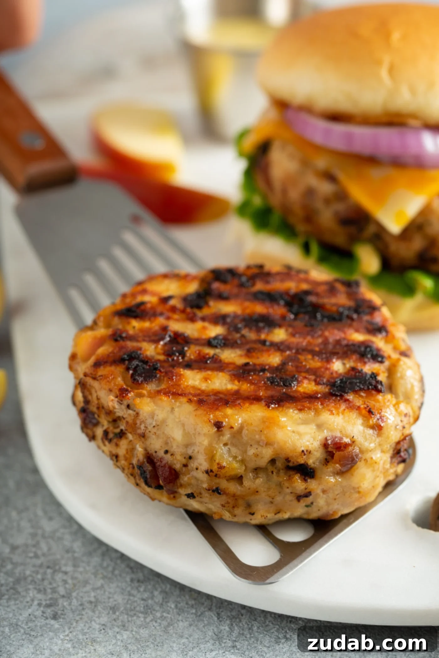 Maple Mustard Apple Chicken Burgers A sizzling, perfectly grilled chicken burger patty being carefully transferred from the grill to a pristine white plate using a spatula. In the softly blurred background, vibrant apple slices and another succulent chicken burger hint at the delicious meal to come.