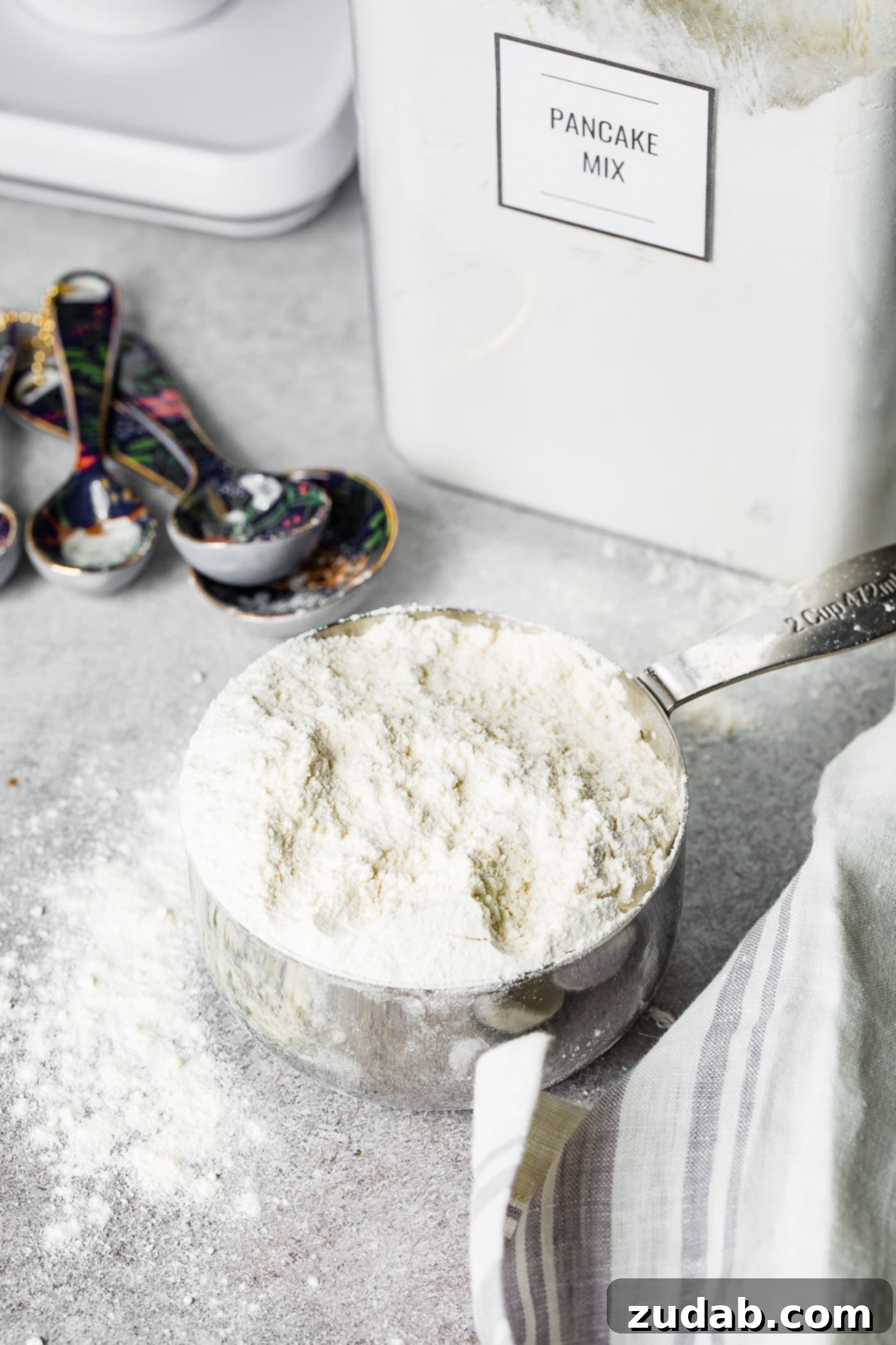 Easy Homemade Pancake Mix Recipe 4 A metal cup heaping full of homemade pancake mix. there are metal measuring spoons and a clear canister of buttermilk pancake mix in the background.