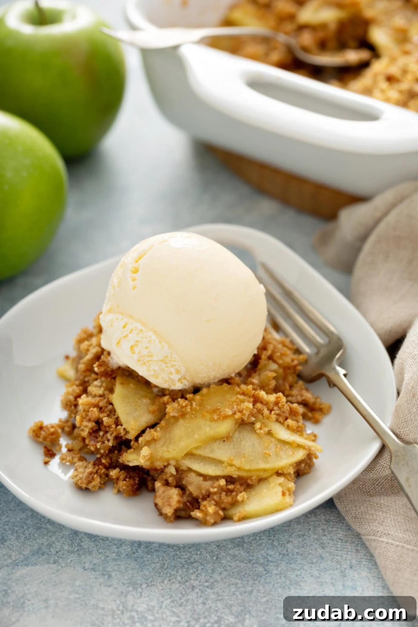 Apple Brown Betty A warm Apple Brown Betty dessert in a dish, topped with a scoop of melting vanilla ice cream and a fork on the plate. Sliced apples and a linen napkin are in the background, hinting at fresh ingredients and homely comfort.