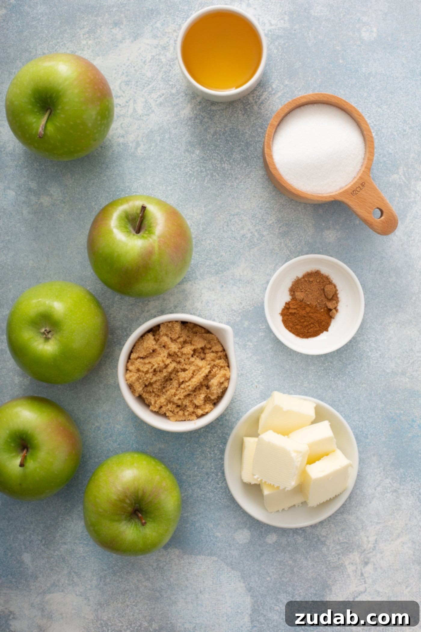 Apple Brown Betty A vibrant selection of five green apples, alongside containers of cubed butter, aromatic cinnamon, crunchy bread crumbs, and sparkling sugar, showcasing the fresh ingredients for an Apple Brown Betty filling.