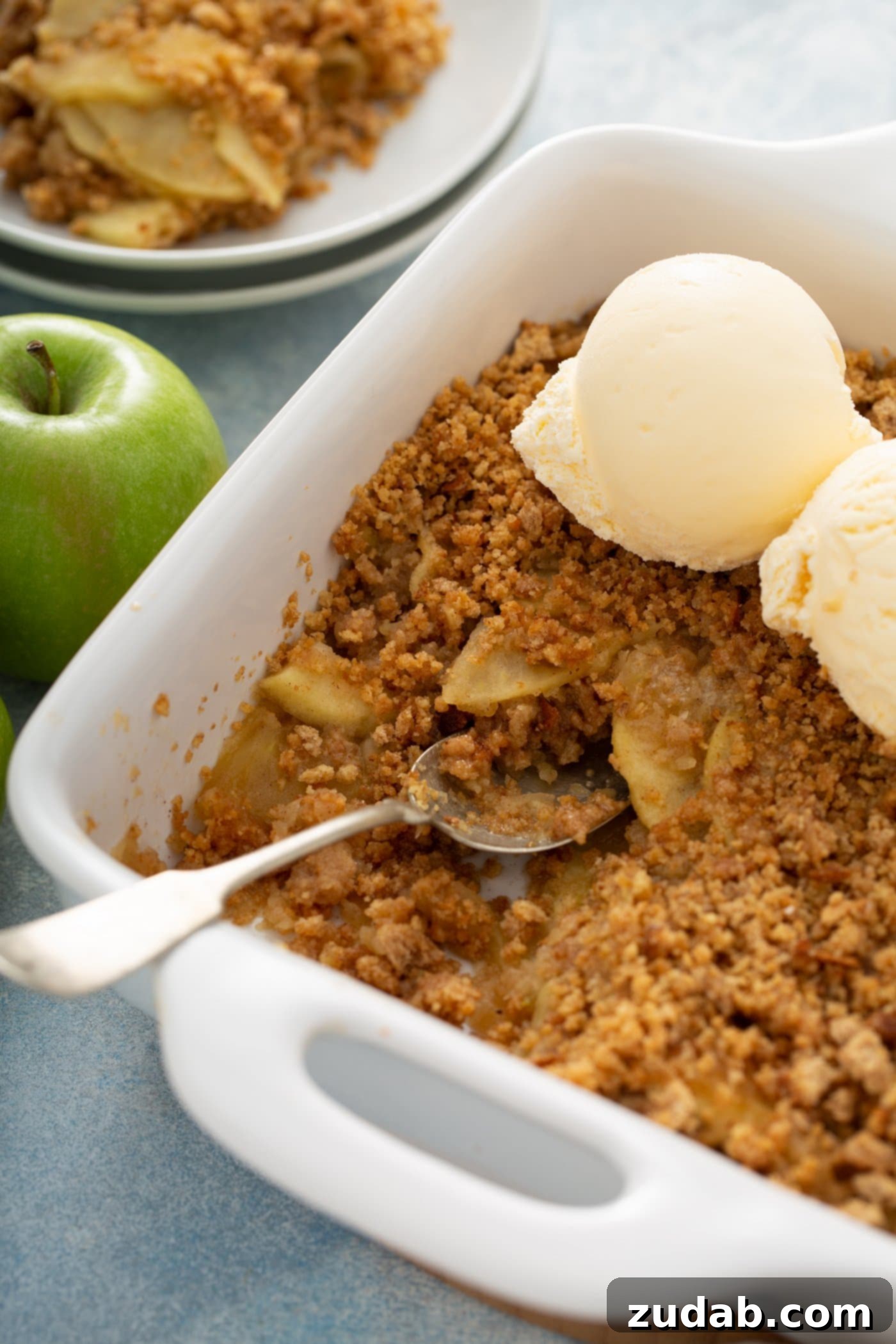 Apple Brown Betty A full baking dish of Apple Brown Betty, crowned with two generous scoops of vanilla ice cream, and a fresh apple on the side. Dessert plates are blurred in the background, signaling a delightful serving.