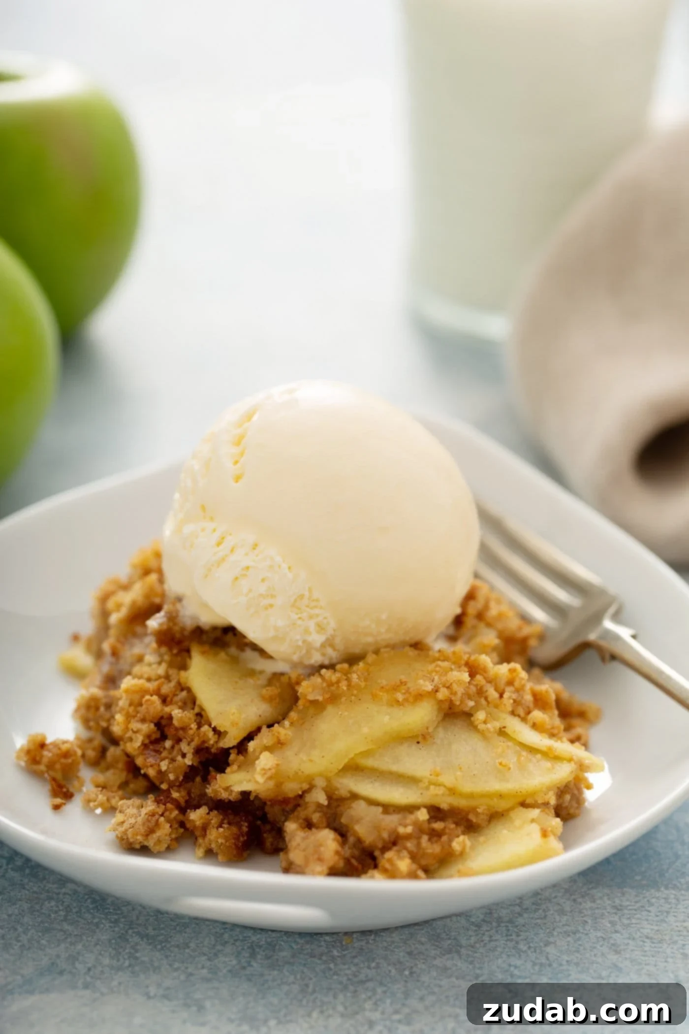 Apple Brown Betty A single serving of Apple Brown Betty on a dessert plate, featuring layered spiced apples with a golden bread crumb topping, finished with a scoop of vanilla ice cream and a fork. Green apples are visible in the soft background.