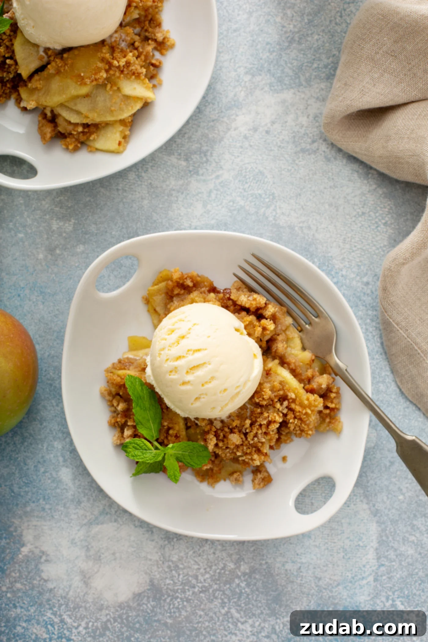Apple Brown Betty A generous serving of Apple Brown Betty on a white dessert plate, adorned with a scoop of vanilla ice cream and a sprig of fresh peppermint, with another serving visible in the soft background.