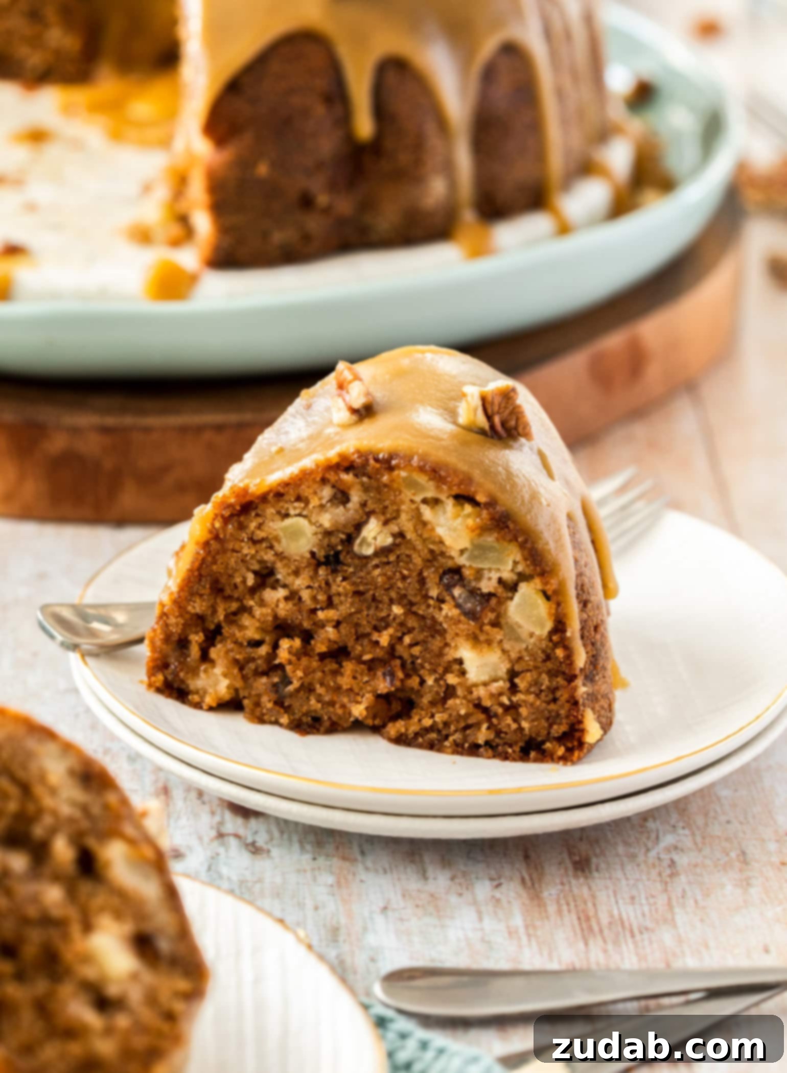 Spiced Apple Pecan Bundt 3 A slice of apple bundt cake sitting on a white plate. The rest of the cake is in the background