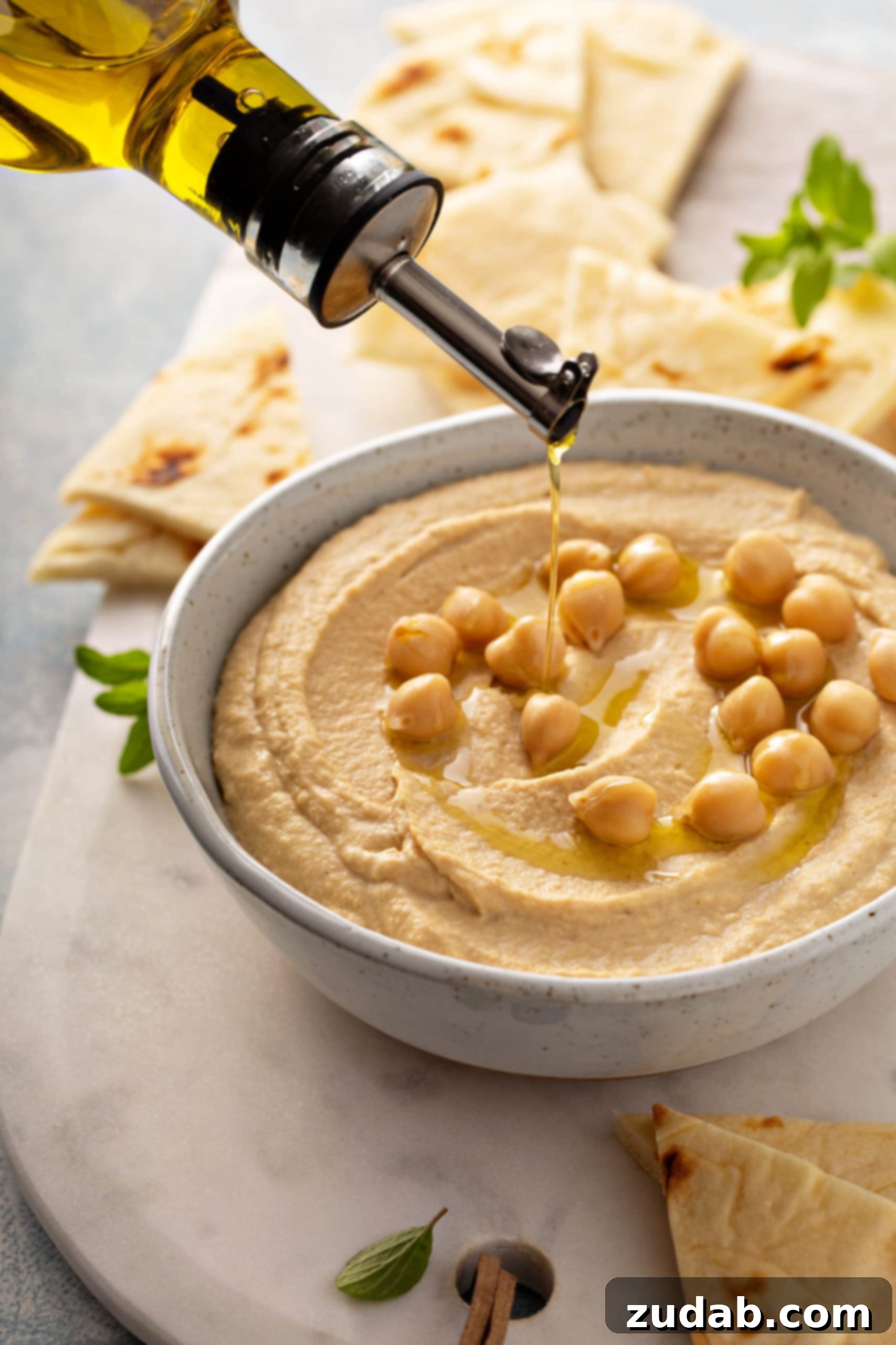 Drizzling Olive Oil on Hummus Olive oil being drizzled over a freshly prepared bowl of homemade hummus, garnished with whole chickpeas and tortilla wedges in the background.