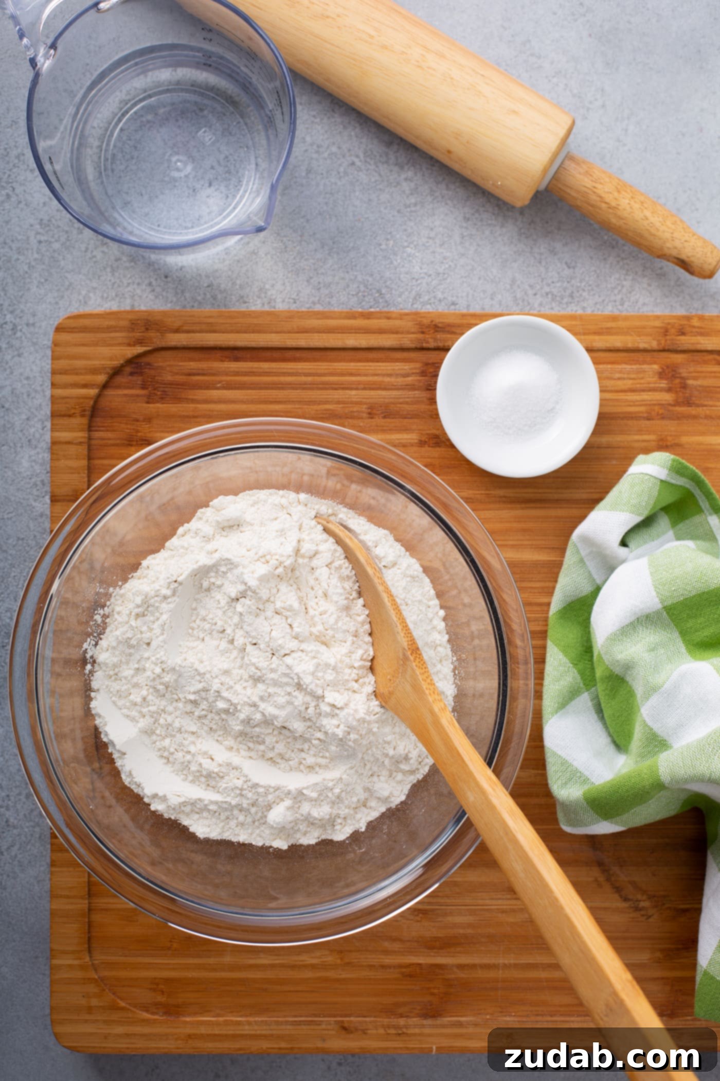 Unleavened Bread A photo of a glass bowl of flour with a wooden spoon in it and a small white bowl of salt sitting on a wooden cutting board with a glass measuring cup of water and wooden rolling pin on the side..